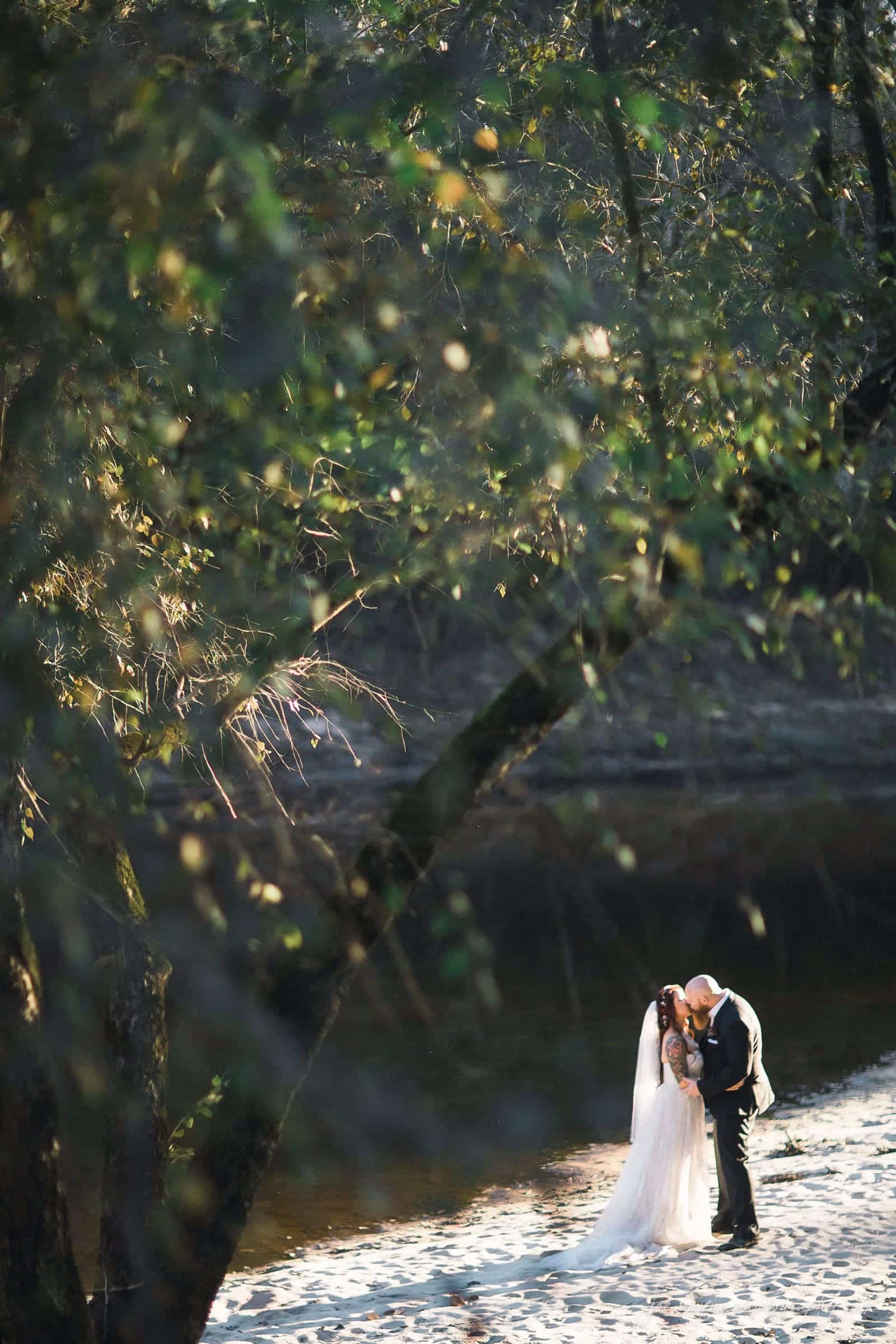 river landing country club wedding - megan & chad A bride and groom share an intimate moment on a sandy riverbank. They are surrounded by lush, green trees with sunlight filtering through the leaves, creating a serene and romantic atmosphere.