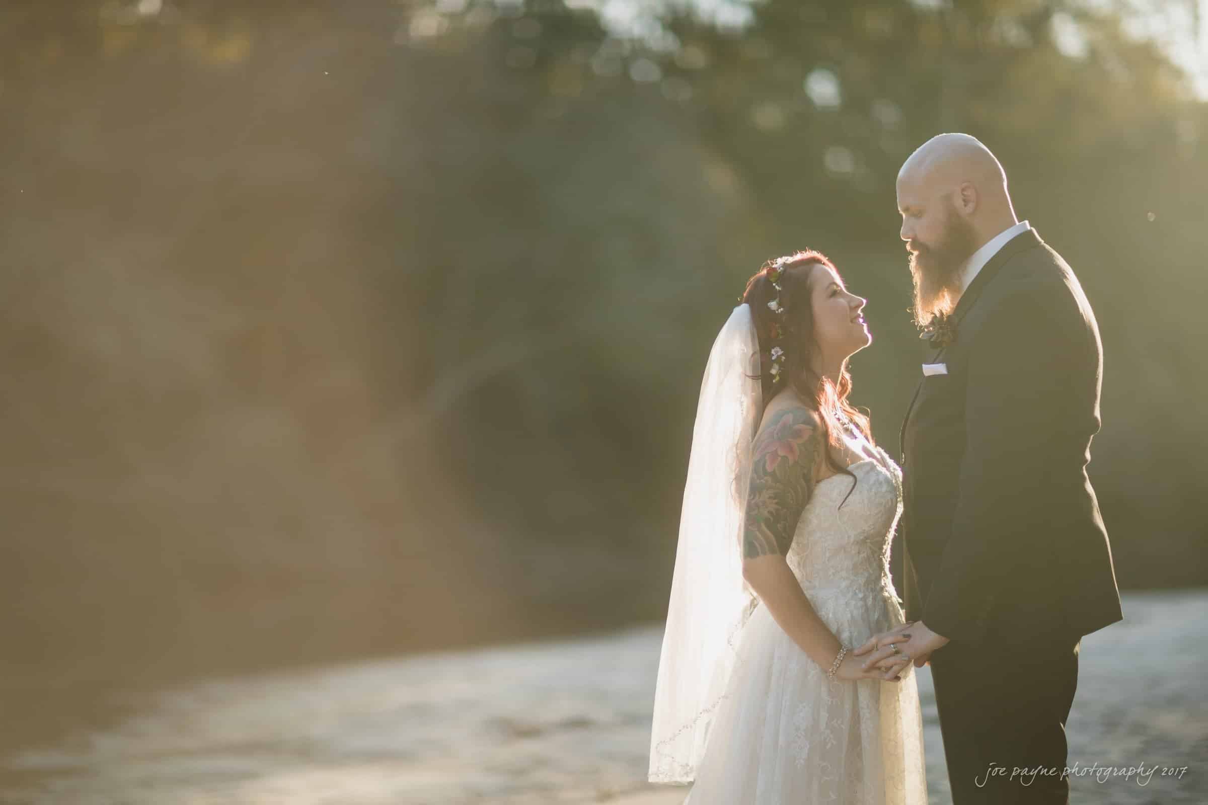 river landing country club wedding - megan & chad A bride and groom stand outdoors, holding hands and facing each other. The bride wears a white dress and veil, while the groom is in a dark suit. Sunlight creates a warm glow around them, with a blurred natural background.
