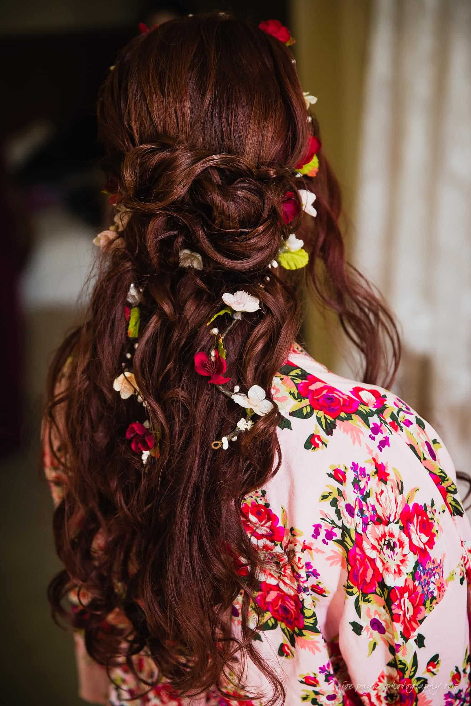 river landing country club wedding - megan & chad A woman with long, wavy brunette hair is adorned with red, white, and green flowers. She wears a floral-patterned robe in shades of pink, red, and green. Her back is turned, showing intricate hair styling.