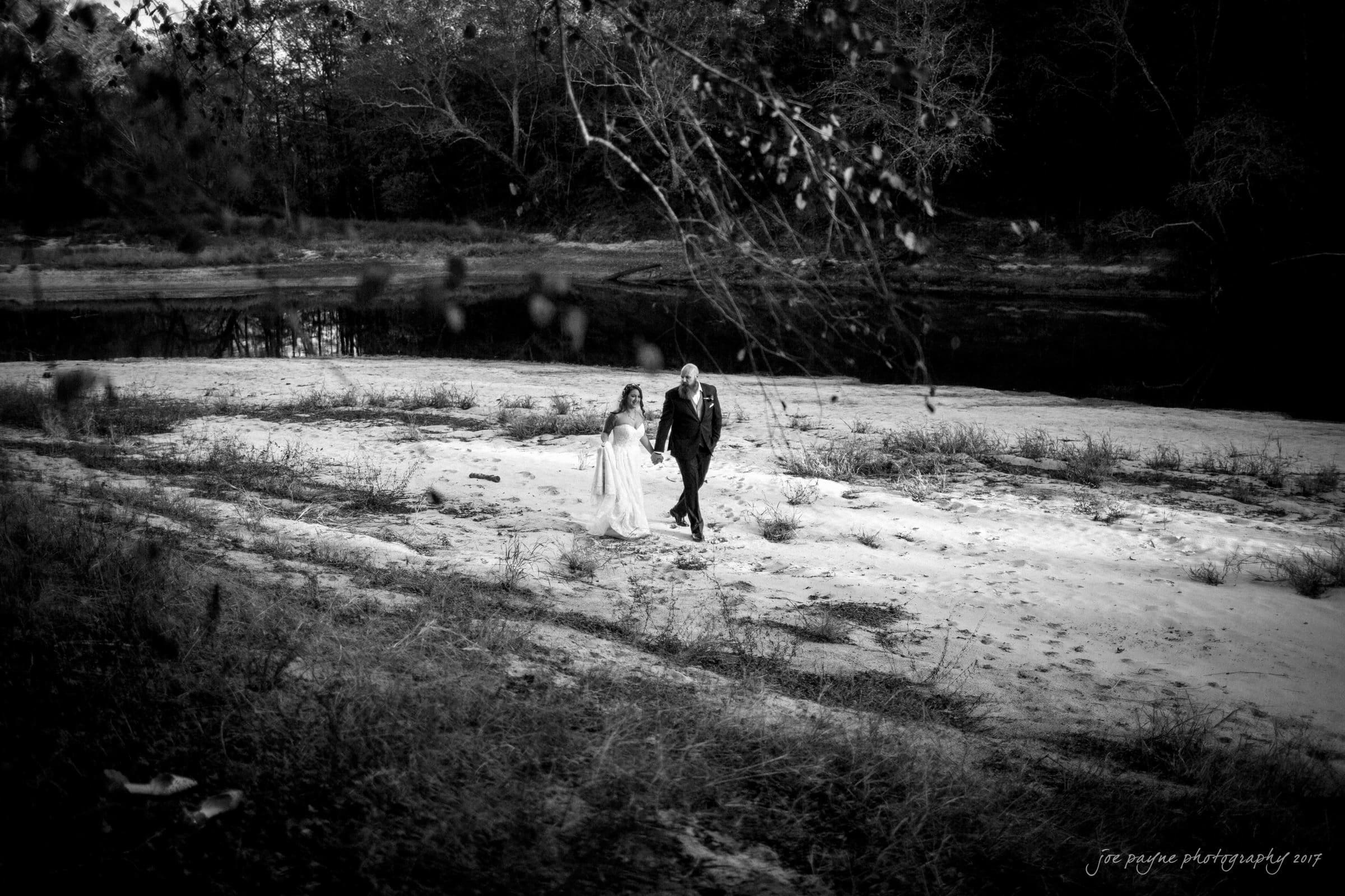 river landing country club wedding - megan & chad A couple in formal attire walks hand in hand on a sandy shore by a calm river. The background features bare trees and a serene, natural setting, captured in black and white.