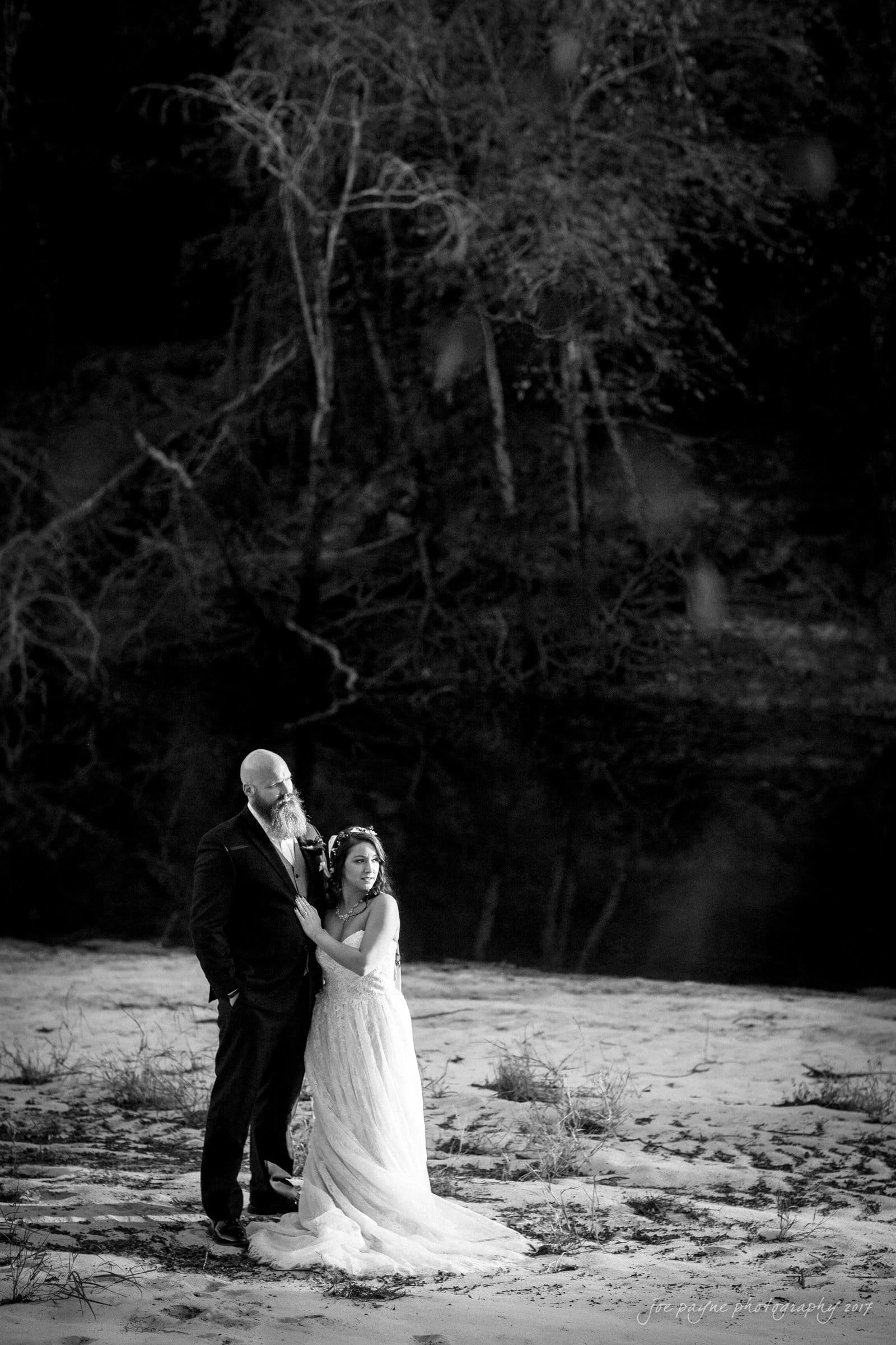 river landing country club wedding - megan & chad A bride in a flowing gown and a groom in a suit stand together on a sandy riverbank. They are surrounded by trees, with a calm river in the background. The black and white image captures a serene and intimate moment.