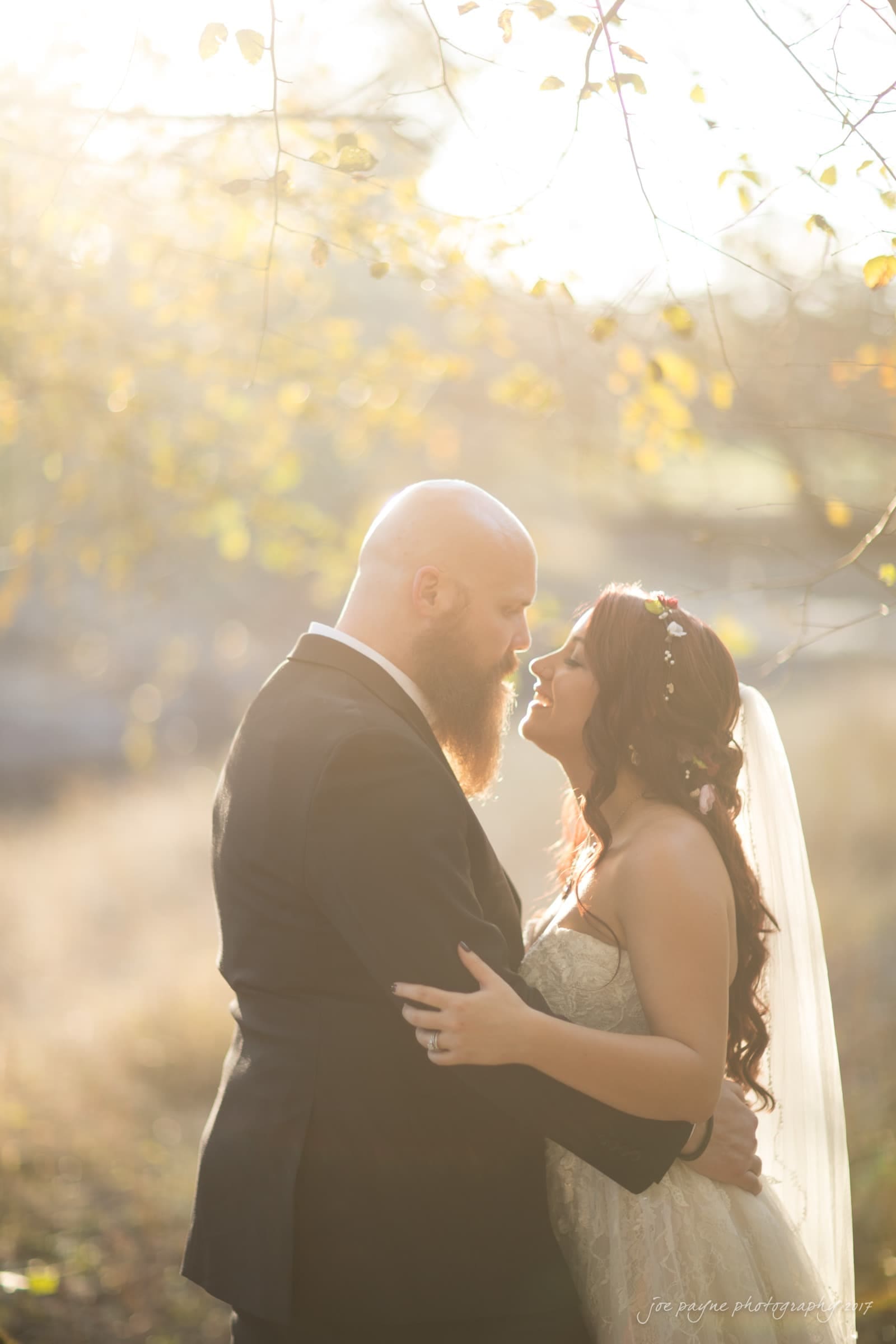 river landing country club wedding - megan & chad A bride and groom stand facing each other, lovingly embracing in a sunlit outdoor setting. The bride wears a veil and floral headpiece, while the groom has a full beard and suit. Sunlight filters through the trees, creating a warm, romantic atmosphere.