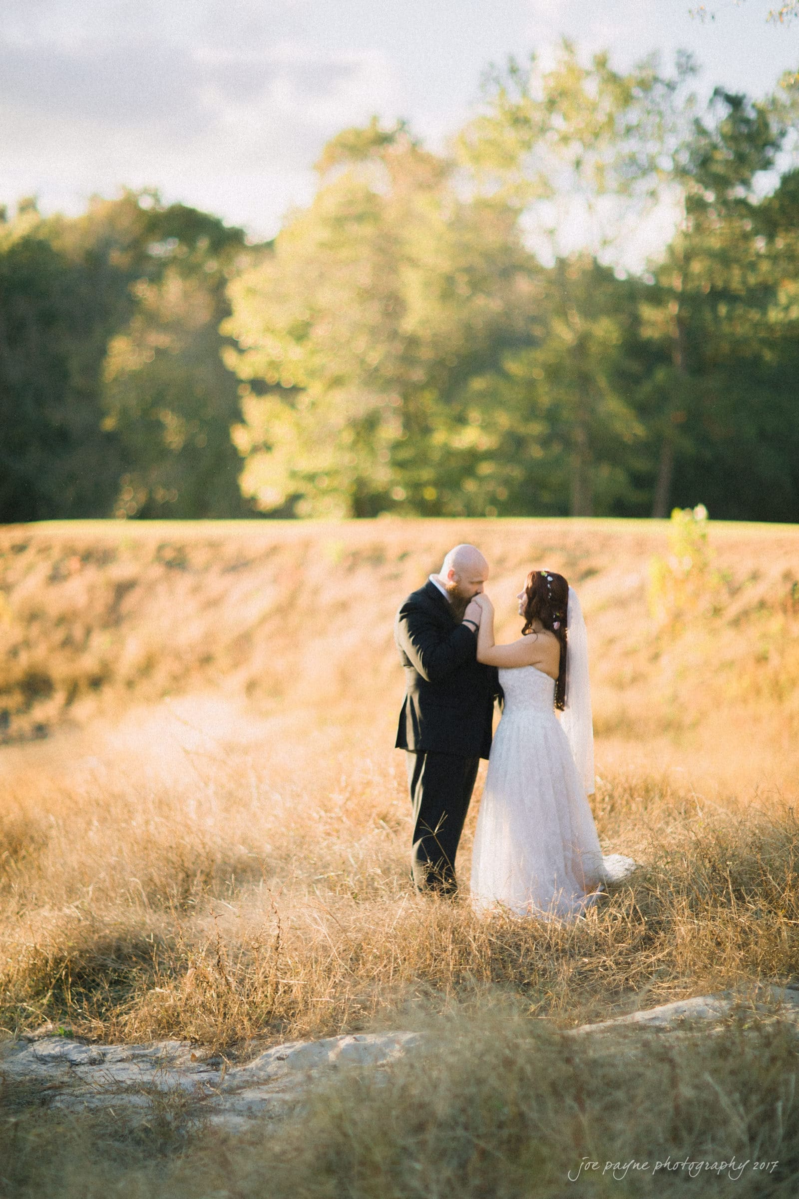 river landing country club wedding - megan & chad A couple stands in a sunlit meadow surrounded by tall grass. The man, in a black suit, gently holds the woman, who is wearing a white wedding dress. Trees and a clear sky form the background, creating a serene, romantic scene.