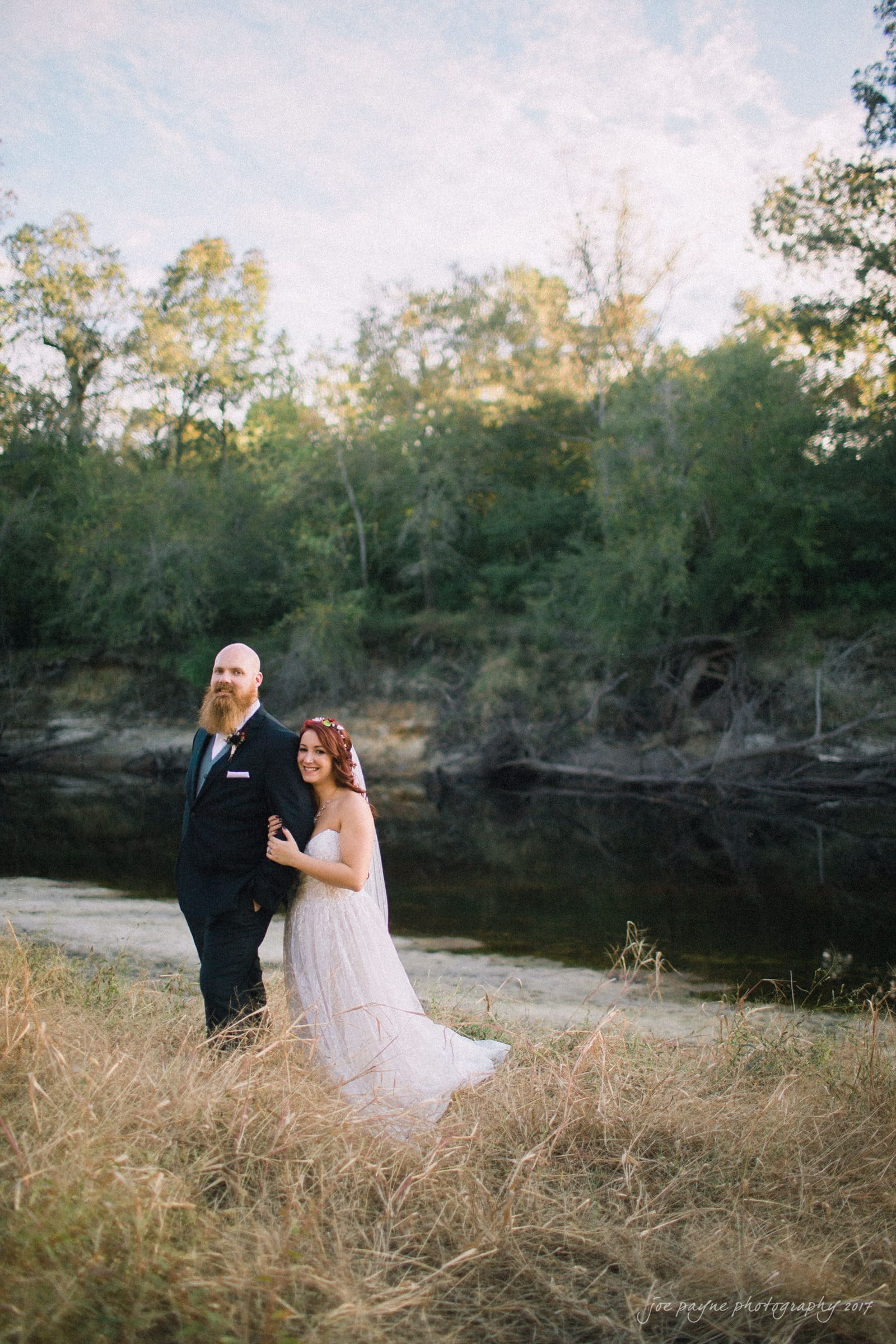 river landing country club wedding - megan & chad A couple is walking hand in hand outdoors by a river. The groom wears a black suit and tie, the bride wears a white dress and veil. Theyre surrounded by tall grass and trees under a clear sky.