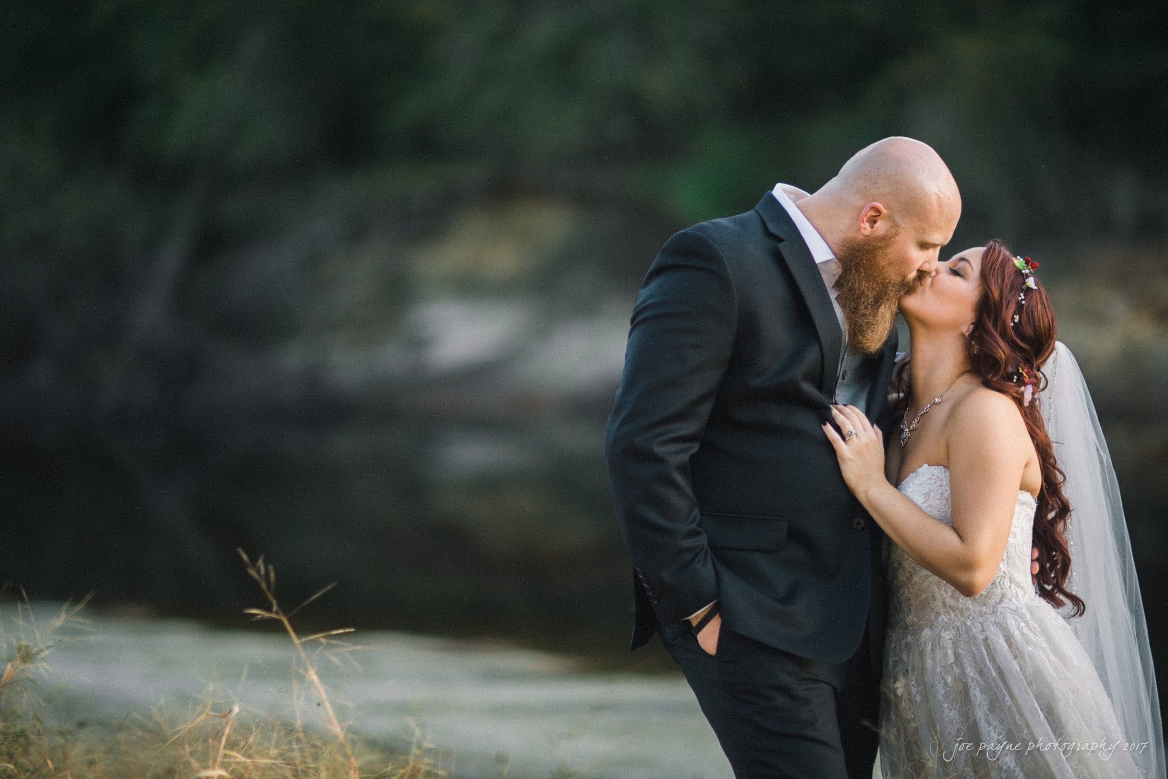 river landing country club wedding - megan & chad A newlywed couple shares a kiss by a serene, wooded riverside. The groom, in a dark suit, holds the bride close. Shes wearing a white wedding dress with a veil and floral crown. The background is softly focused with greenery and a calm river.