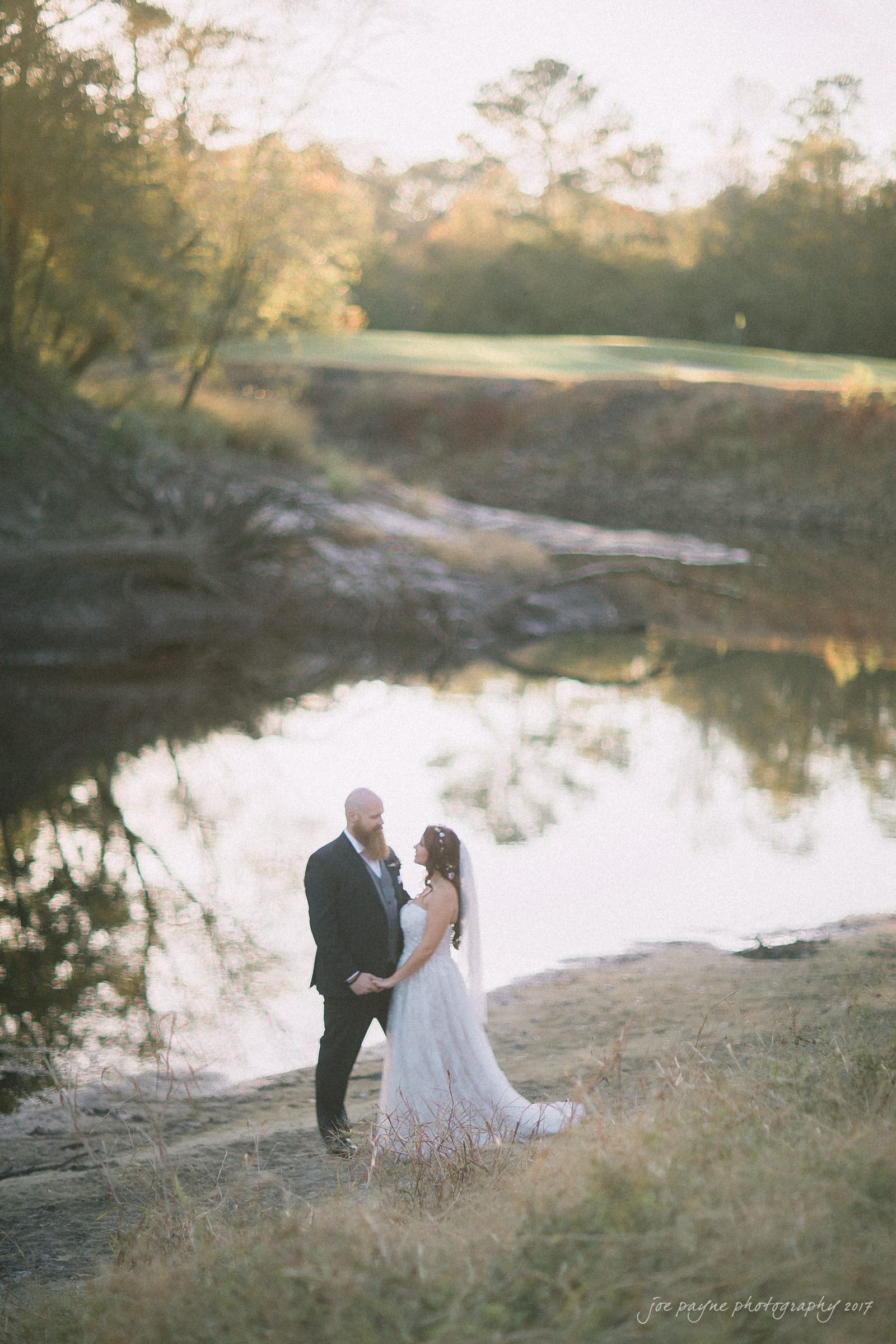 river landing country club wedding - megan & chad A couple in wedding attire stands beside a tranquil lakeside, surrounded by autumn trees. The bride wears a long white dress and veil, holding hands with the groom in a dark suit. Soft, natural light creates a serene atmosphere.