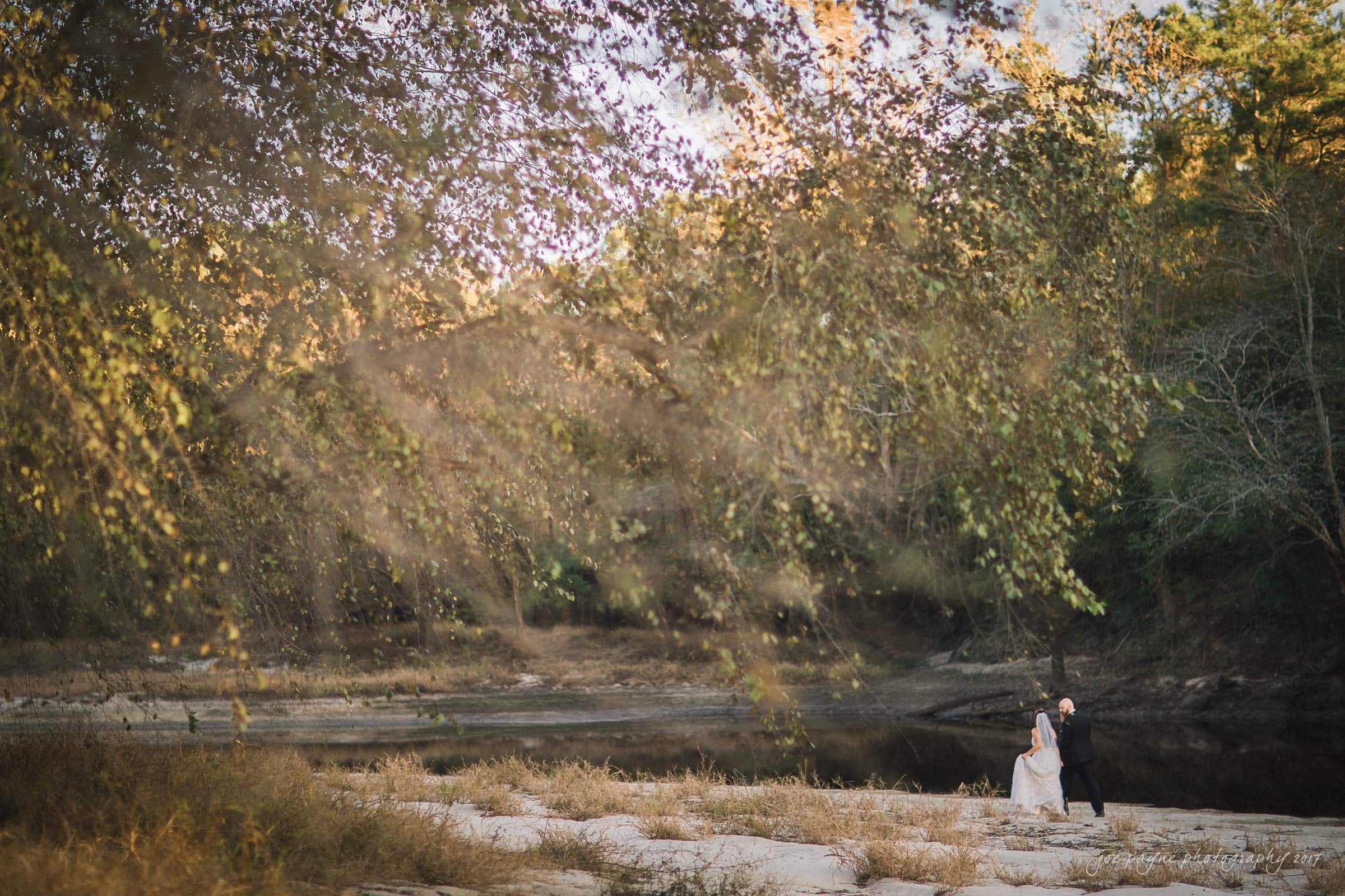 river landing country club wedding - megan & chad A couple in wedding attire walks hand in hand by a serene riverside, surrounded by tall trees with autumn leaves. The scene is peaceful, with the natural beauty of the landscape creating a romantic ambiance.