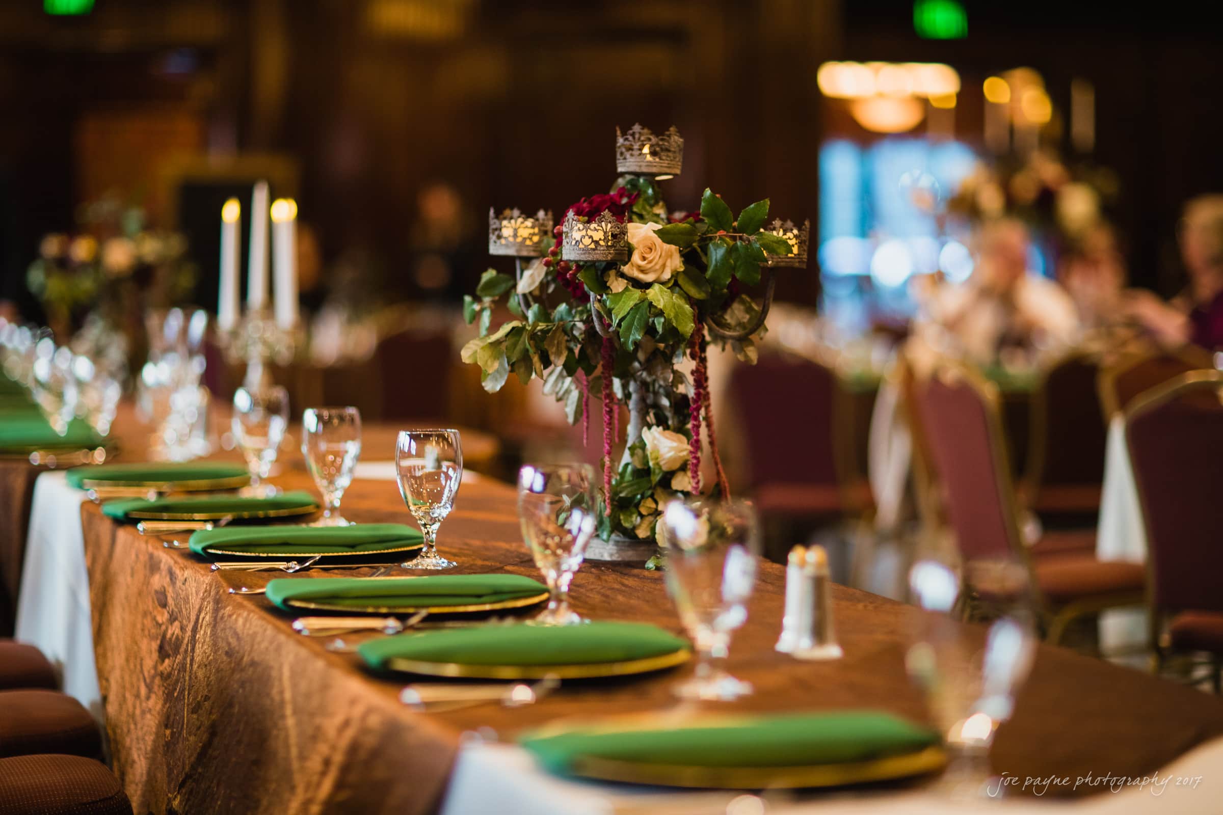 river landing country club wedding - megan & chad A beautifully set banquet table with green napkins, elegant floral centerpieces with crowns, and tall candles. Chairs line either side, and the warm lighting creates an inviting atmosphere in the dining area.