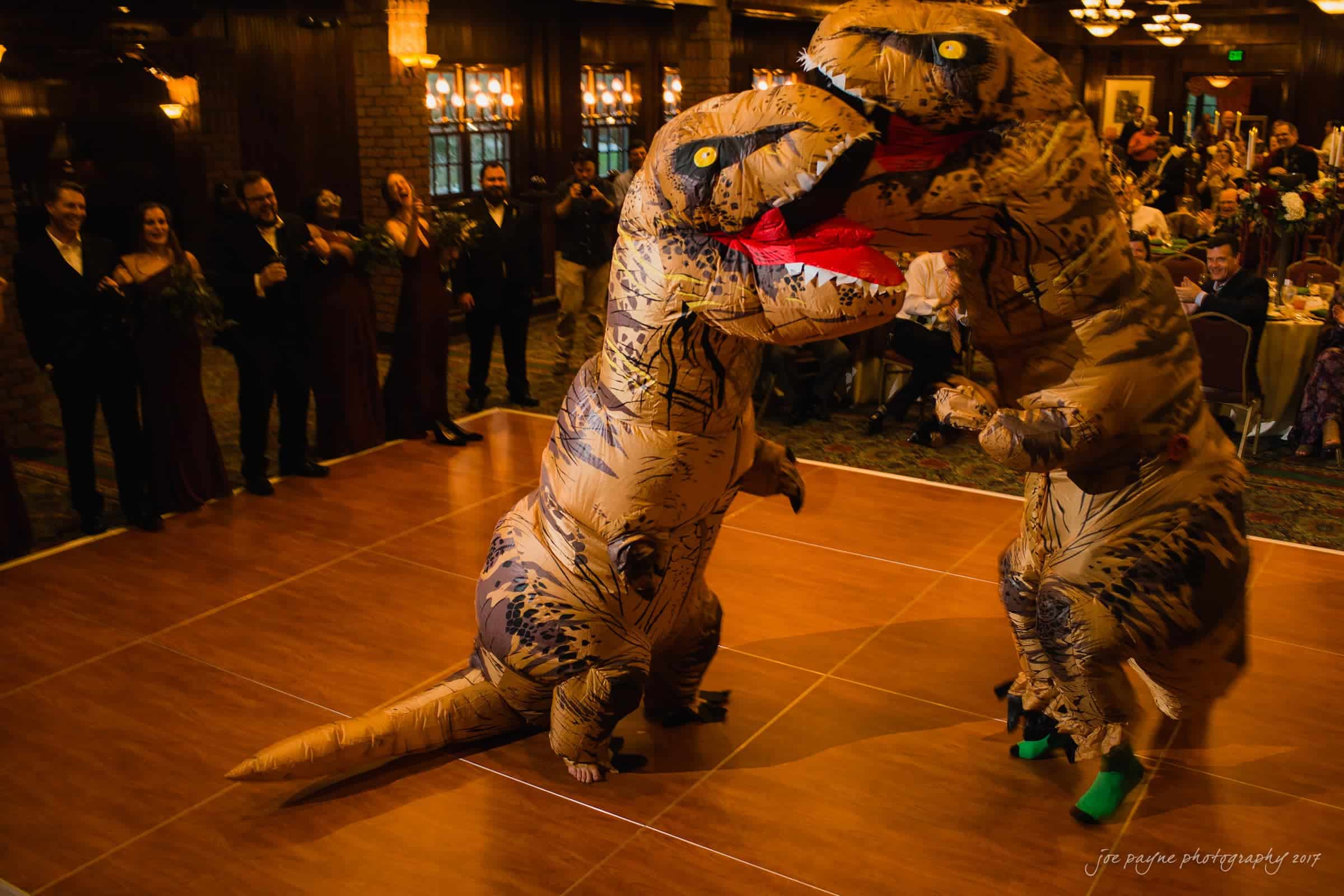river landing country club wedding - megan & chad Two people in inflatable dinosaur costumes dance on a wooden floor at a party. Guests dressed in formal attire watch and laugh in the background. The room is warmly lit and decorated for a festive event.