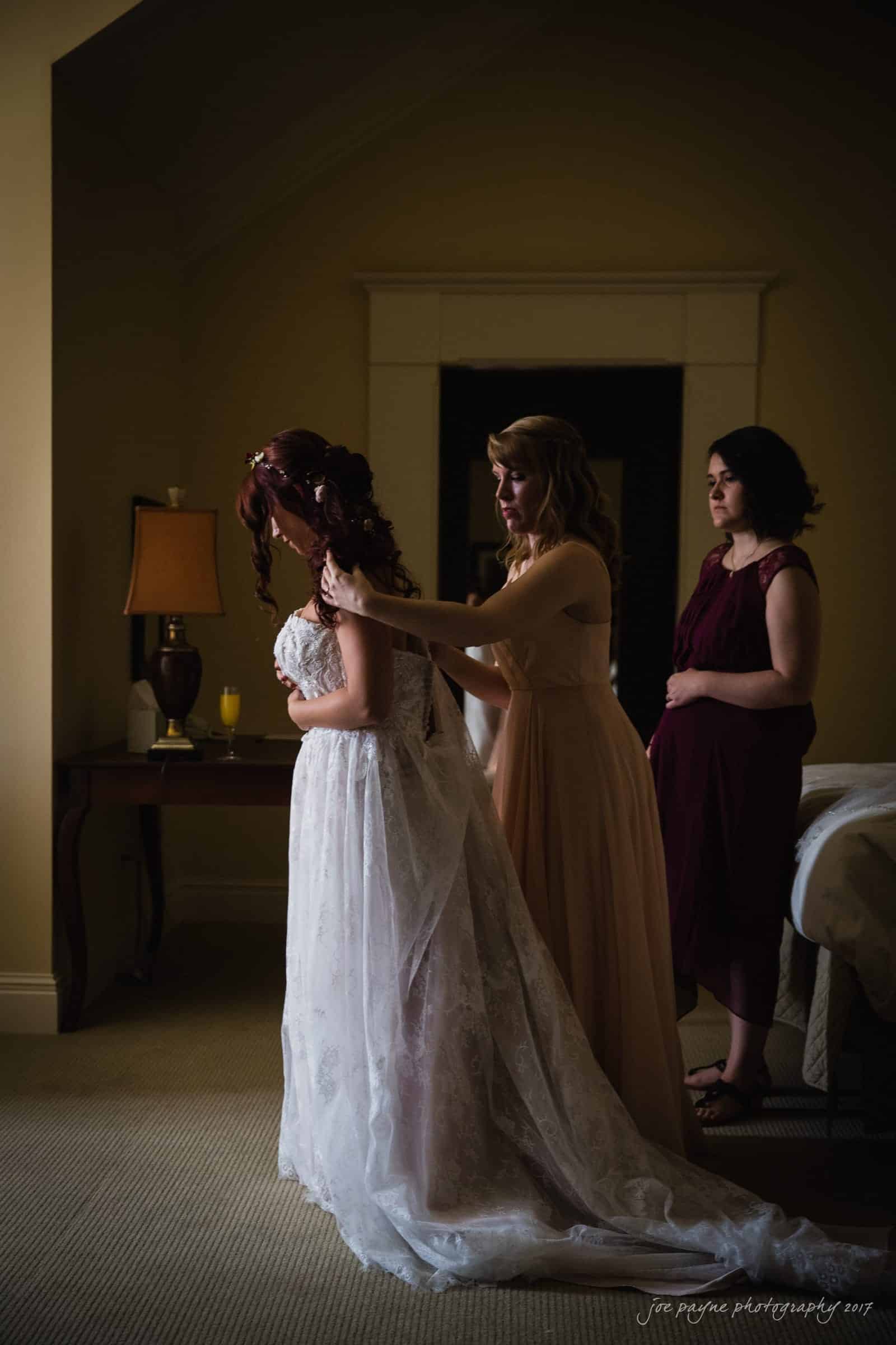 river landing country club wedding - megan & chad A bride in a white lace dress stands in a softly lit room. Two bridesmaids, one in a light dress and one in a dark red dress, assist her with her gown. A bedside lamp and a drink on the table are visible in the background.