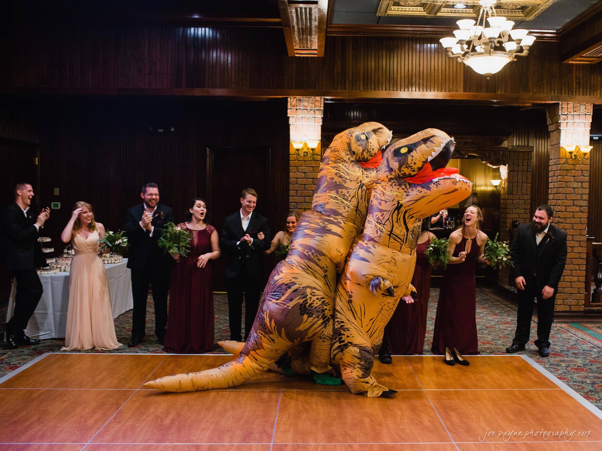 river landing country club wedding - megan & chad Two people in inflatable dinosaur costumes dance together on a wooden floor at a formal event. Guests in evening wear, including bridesmaids and groomsmen, stand around them clapping and smiling in a dimly-lit, elegant room.