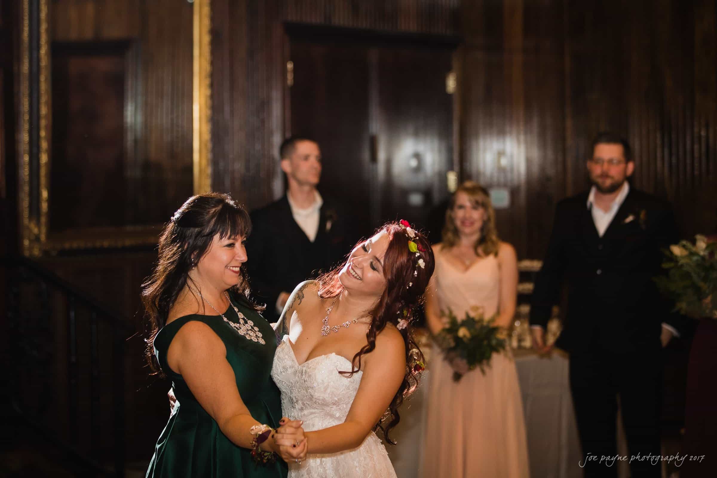 river landing country club wedding - megan & chad A bride in a white dress and floral crown dances joyfully with a woman in a green dress. In the background, three people in formal attire observe with smiles in a warmly lit room.