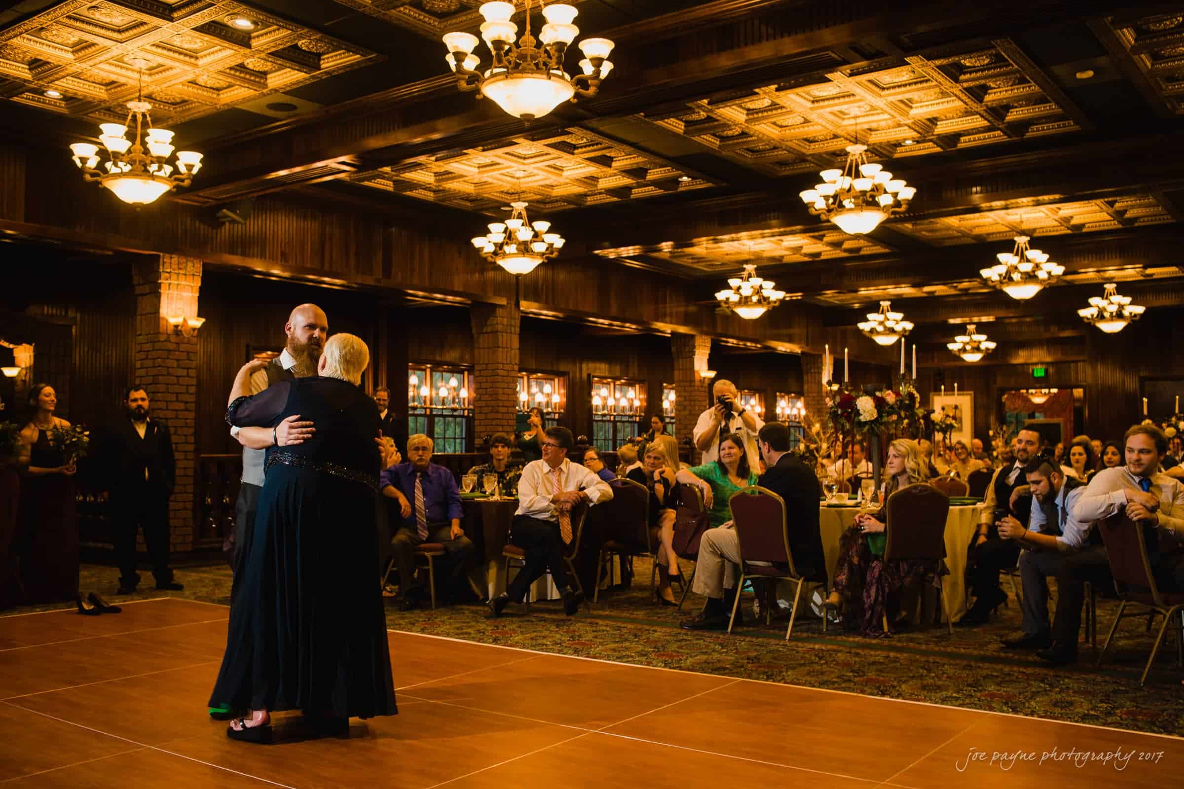 river landing country club wedding - megan & chad A couple dances intimately on a warmly lit ballroom floor, surrounded by seated guests at round tables. Decorative lights and ornate ceiling tiles add elegance to the scene.