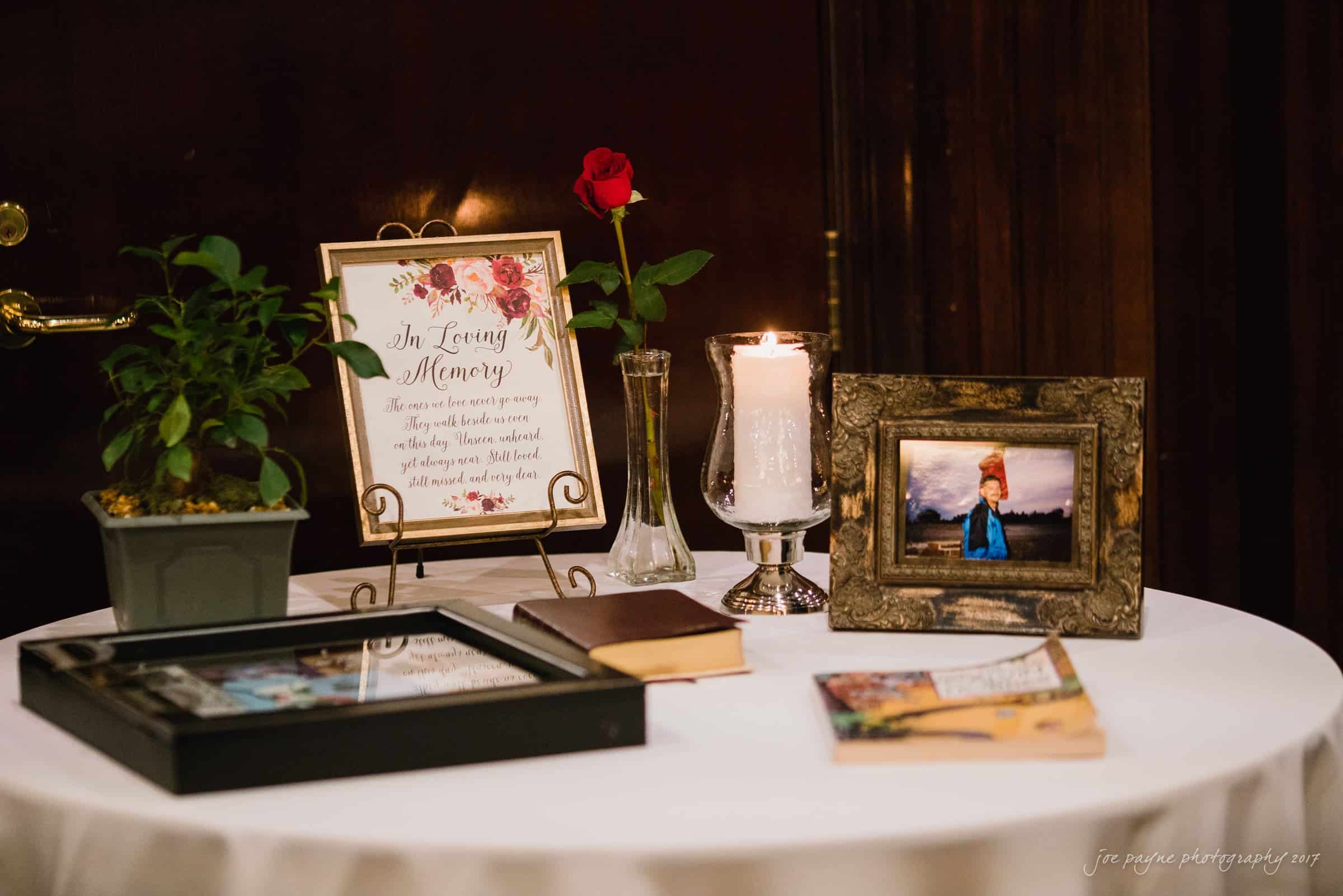 river landing country club wedding - megan & chad A table memorial display with a framed quote, a single red rose in a vase, a lit candle, and framed photos. A small potted plant and books are also arranged on the white tablecloth.