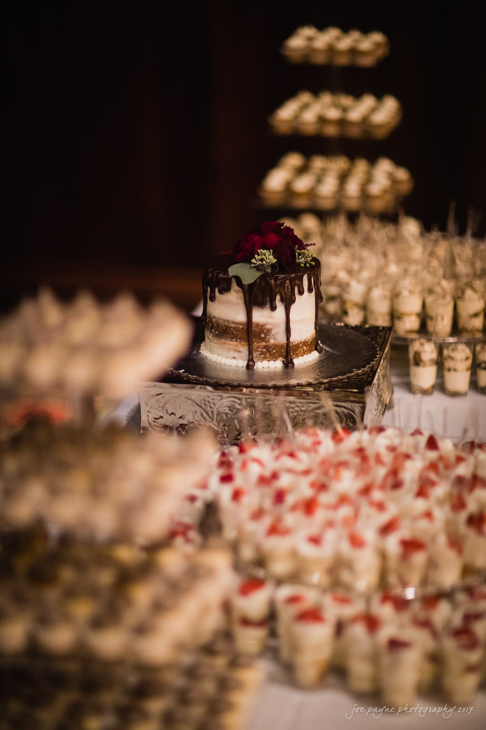 river landing country club wedding - megan & chad A decorated cake with chocolate drips and red flowers on top sits on a square stand. Surrounding it are multiple tiers of dessert cups, possibly with layers of cream and fruit, arranged on a white tablecloth.