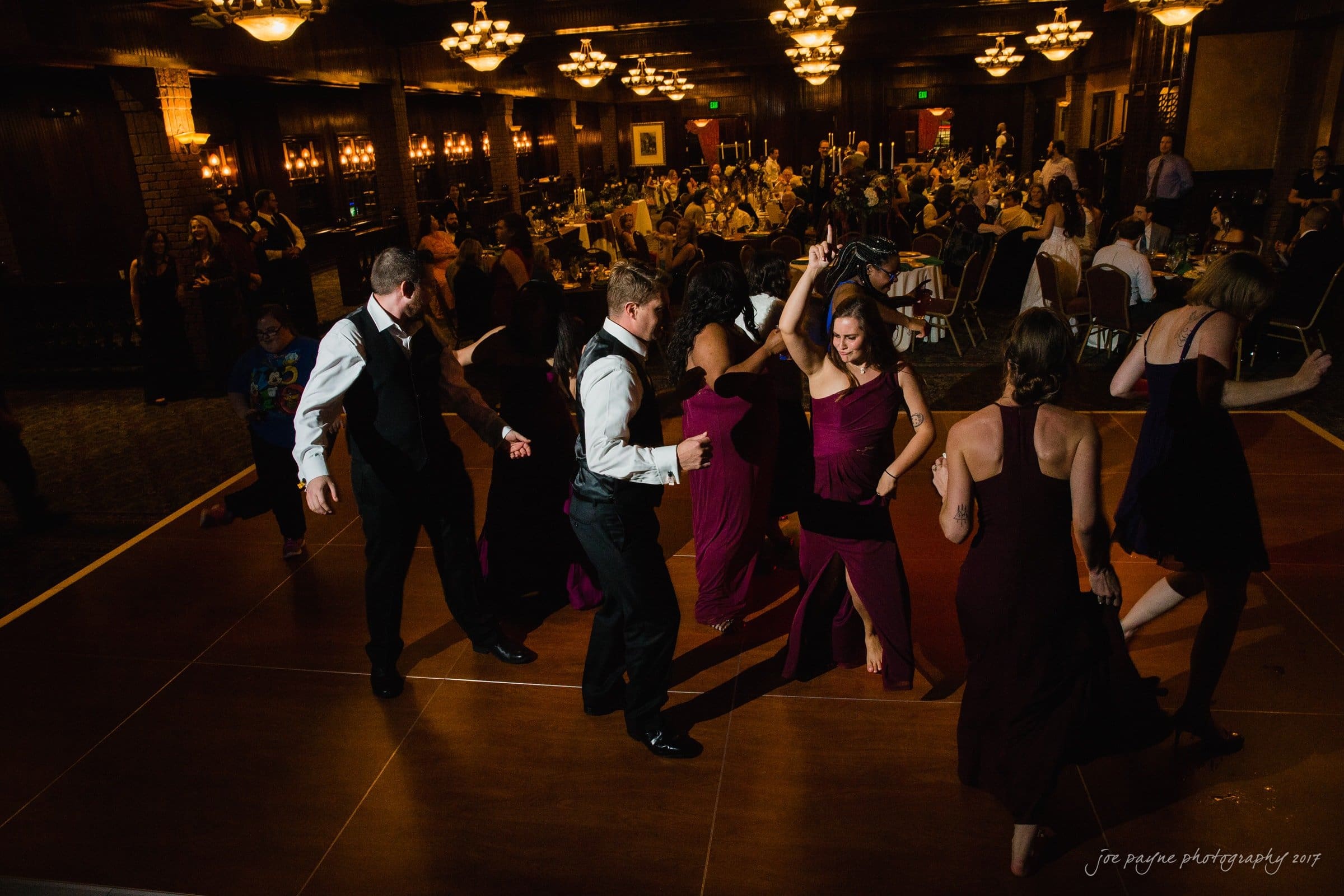river landing country club wedding - megan & chad A group of people, dressed in formal wear, dance energetically on a wooden dance floor at an indoor event. The room is warmly lit, with many seated guests in the background.