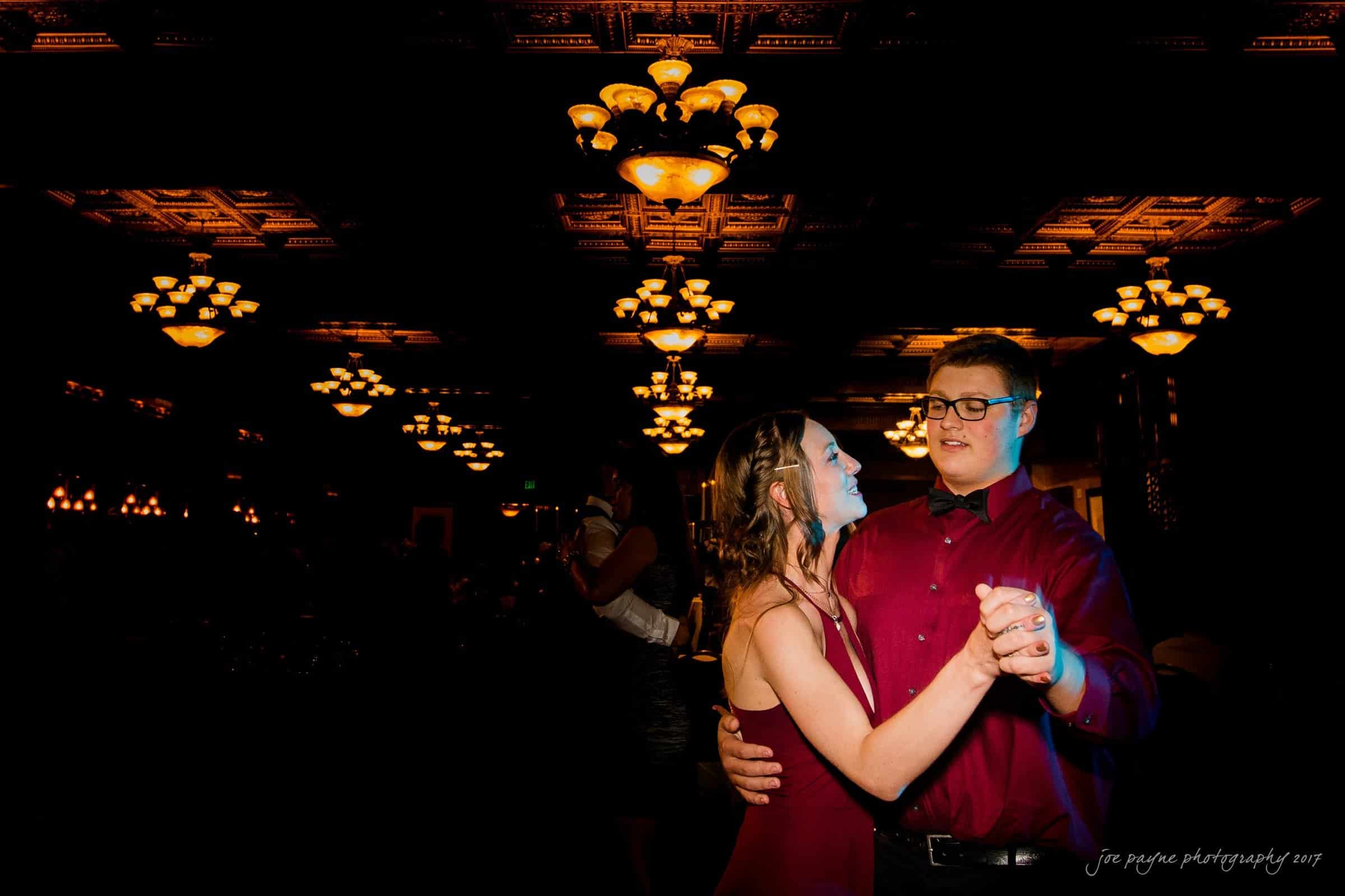 river landing country club wedding - megan & chad A couple dressed in red dances together in a dimly lit room adorned with elegant chandeliers. The man wears glasses and a shirt, while the woman wears a sleeveless dress. They are smiling and sharing a joyful moment.