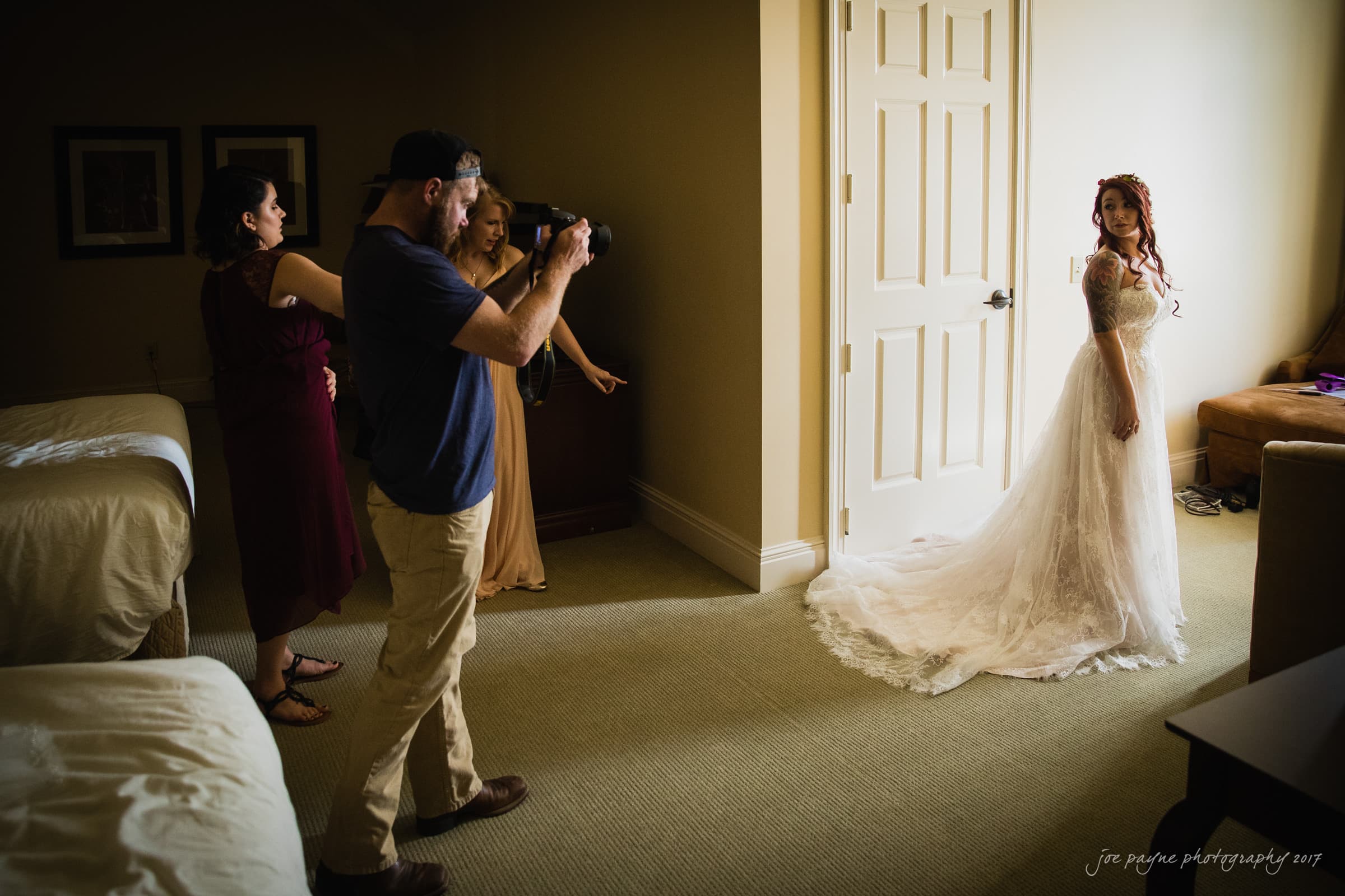 river landing country club wedding - megan & chad A bride in a white gown stands near a door as a photographer takes her picture in a dimly lit room. Two women assist with her dress and styling. The room has simple decor and warm lighting.