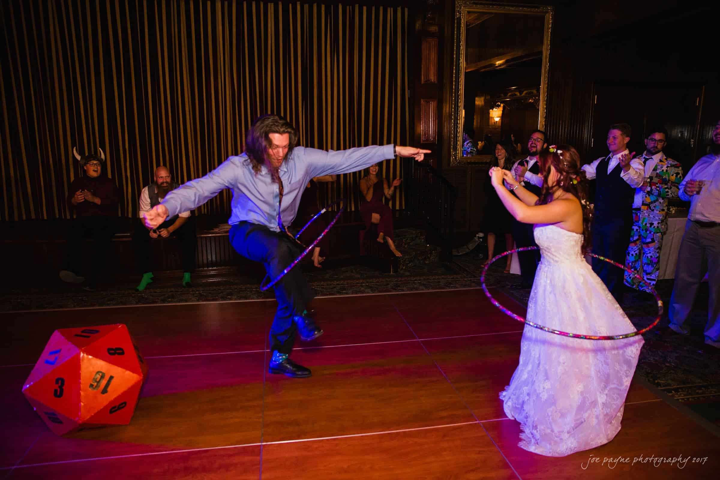 river landing country club wedding - megan & chad A bride and a man joyfully hula hoop on a dance floor. The room features elegant decor and onlookers in the background, including a person in colorful attire. A large 20-sided die is on the floor beside them.