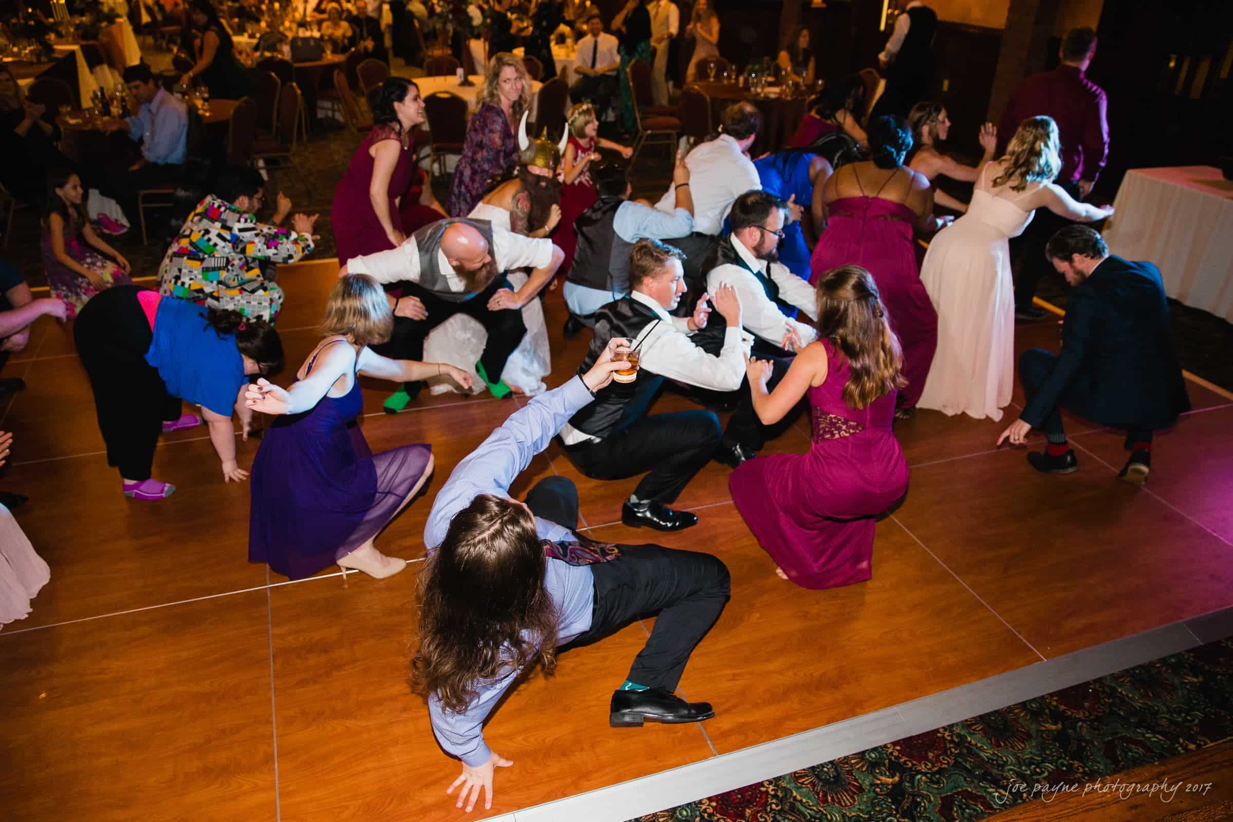river landing country club wedding - megan & chad A lively group of people, dressed in formal attire, dance energetically on a wooden floor at a party or wedding reception. They are bent low, some squatting, in a spirited pose. Tables and other guests are visible in the background.