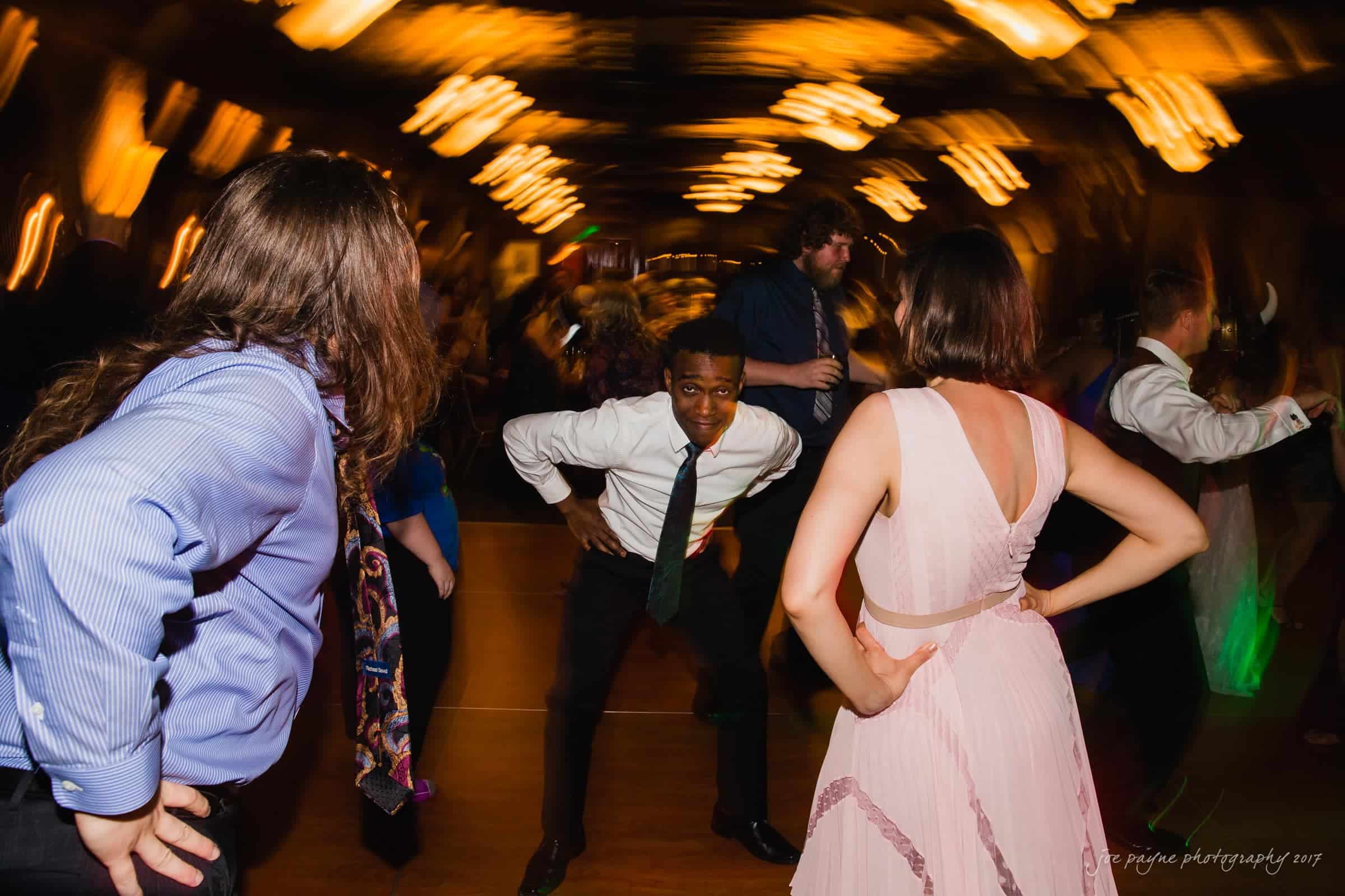 river landing country club wedding - megan & chad A lively dance scene with people dressed in formal attire. A man in the center strikes a playful pose while others around him are dancing energetically. The background features blurred lights, adding a dynamic feel to the atmosphere.