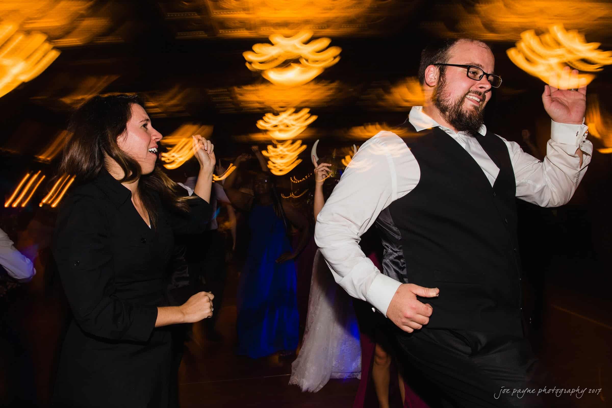 river landing country club wedding - megan & chad A man and woman are dancing energetically at a lively event. The background shows blurred lights resembling chandeliers, creating a festive atmosphere. The man is wearing a vest and glasses, while the woman is dressed in black.