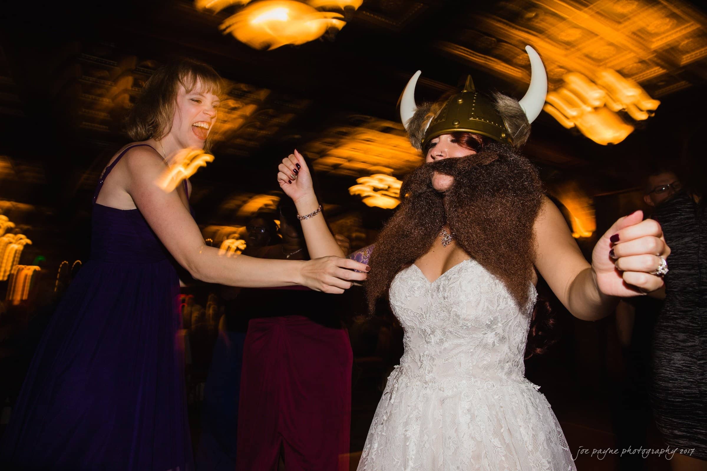 river landing country club wedding - megan & chad A person in a white dress and Viking helmet dances energetically, with a furry beard attached to the helmet. Another person in a purple dress is joyfully dancing nearby. The background is dimly lit with blurred lights.