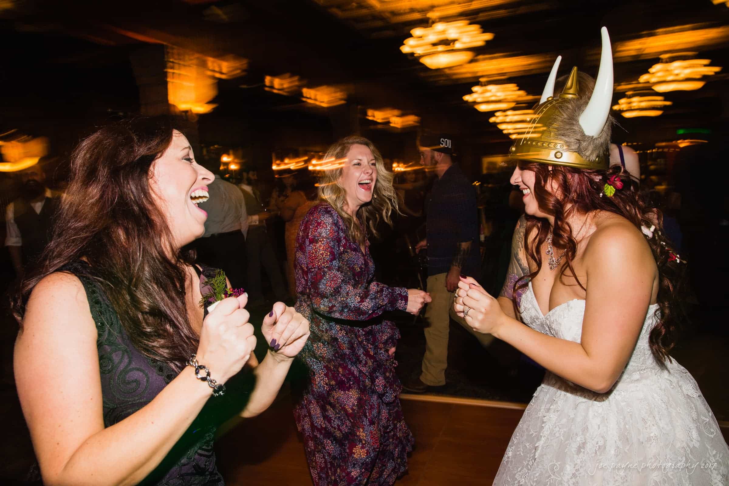 river landing country club wedding - megan & chad Three women are joyfully dancing at a lively indoor event. One woman wears a Viking helmet and a white dress, laughing with the others. The background is blurred, with warm lighting creating a festive atmosphere.