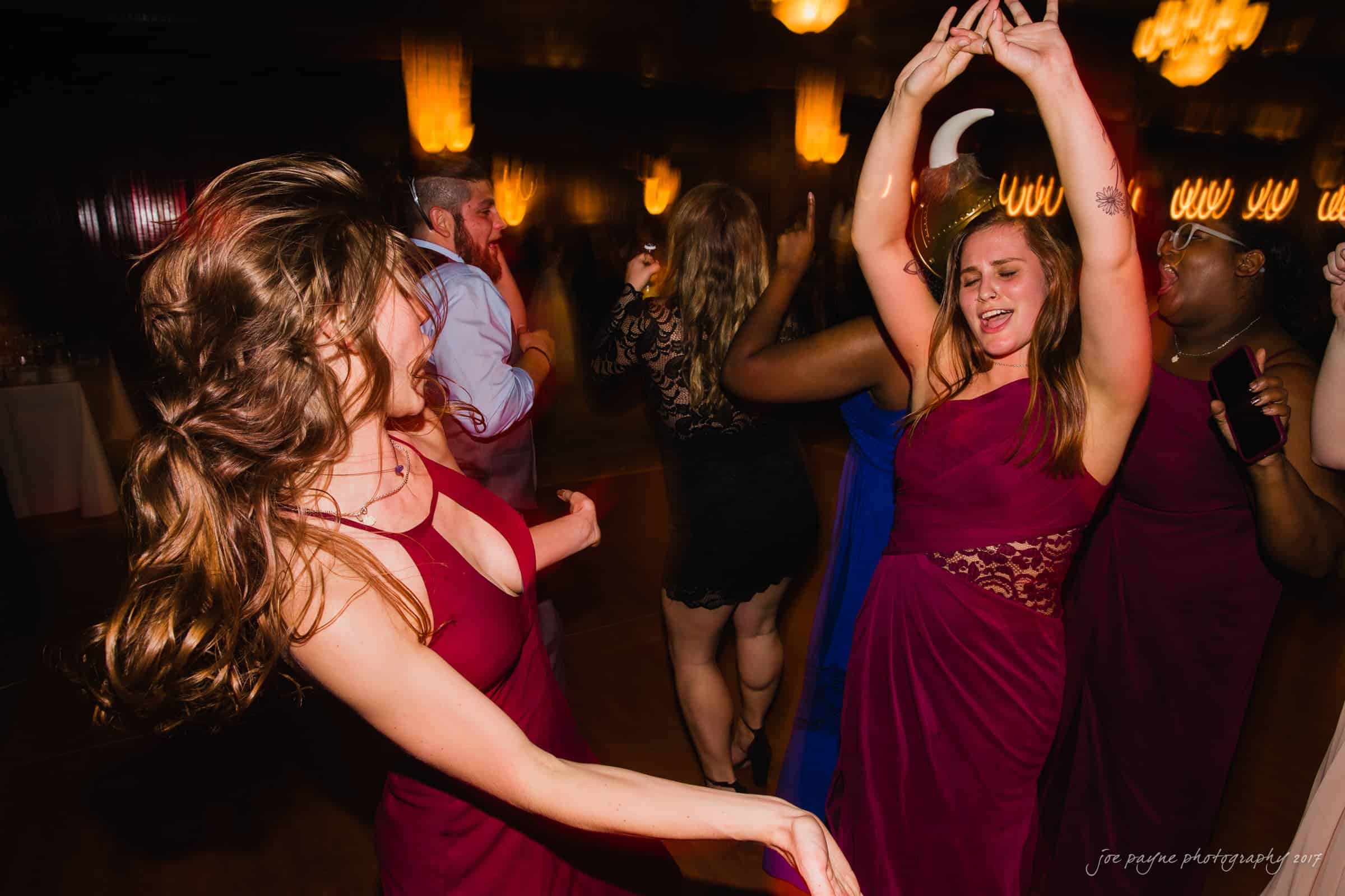 river landing country club wedding - megan & chad A group of people are dancing at a party. Two women in red dresses are prominently enjoying the music, raising their hands. The background is lively, with other guests and warm lighting creating a festive atmosphere.