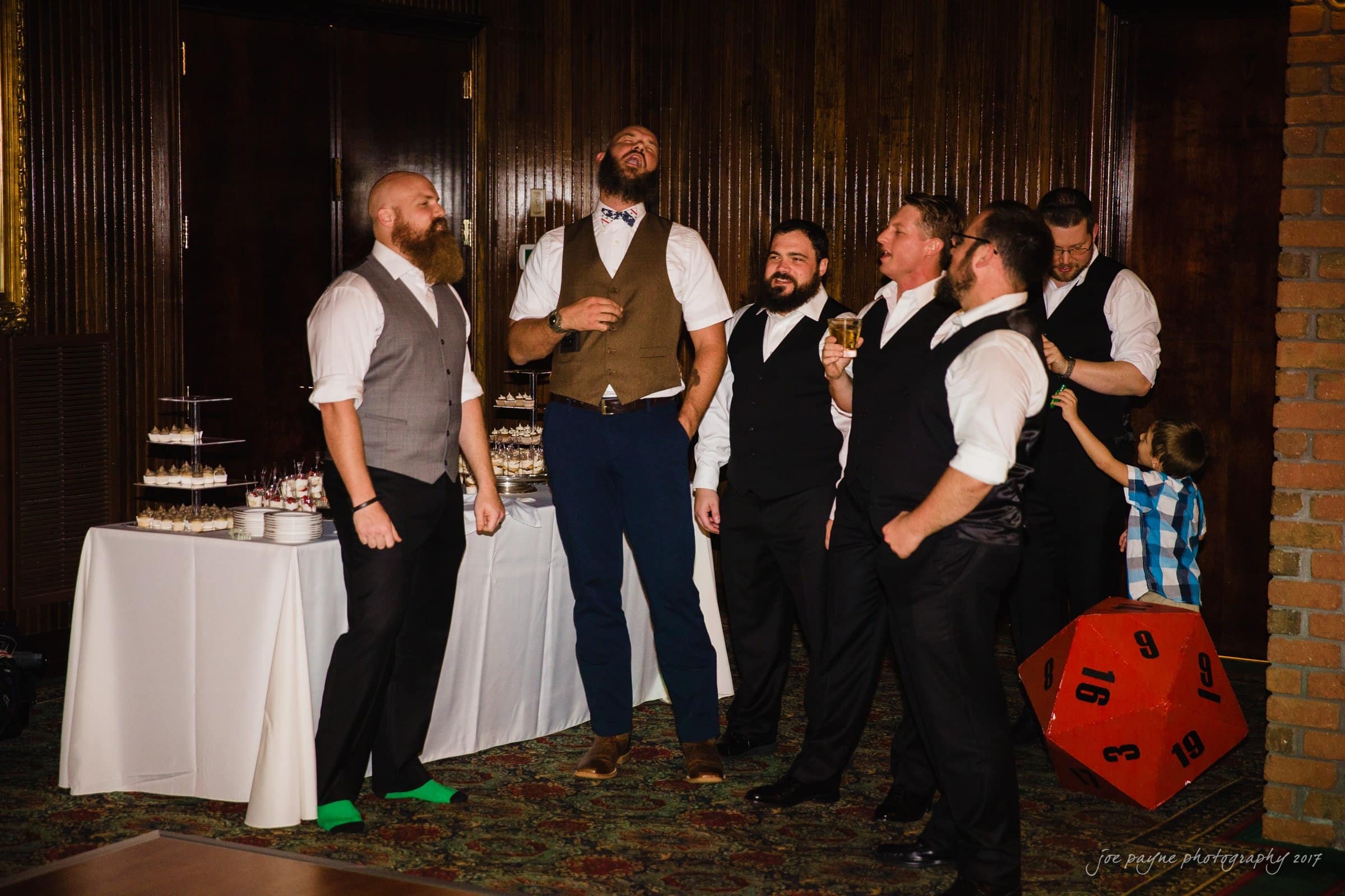 river landing country club wedding - megan & chad A group of six men in formal attire are laughing and talking near a table with cupcakes. One man is looking at a young boy standing beside a large red die. They are in a room with wood-paneled walls and carpeted floors.