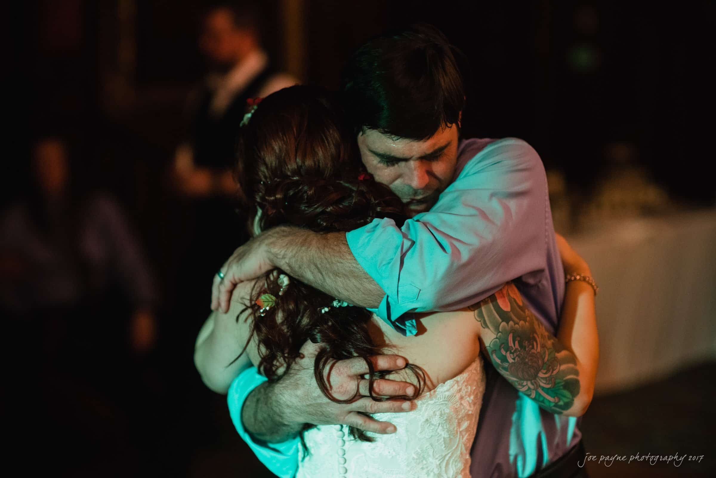 river landing country club wedding - megan & chad A couple embraces closely on a dimly lit dance floor. The woman wears a white dress, and the man has a tattooed arm and is in a purple shirt. They appear to be slow dancing in an intimate moment, surrounded by blurred figures in the background.