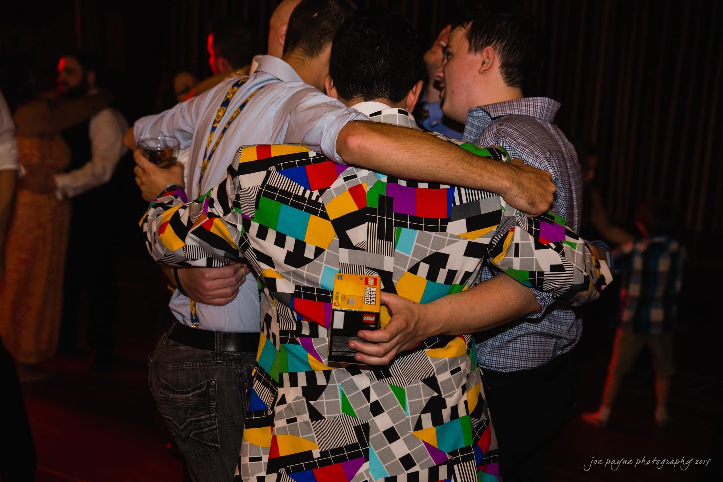 river landing country club wedding - megan & chad A group of people embracing at a party. One person is wearing a colorful, geometric-patterned jacket. The setting is dimly lit, creating a warm and festive atmosphere.