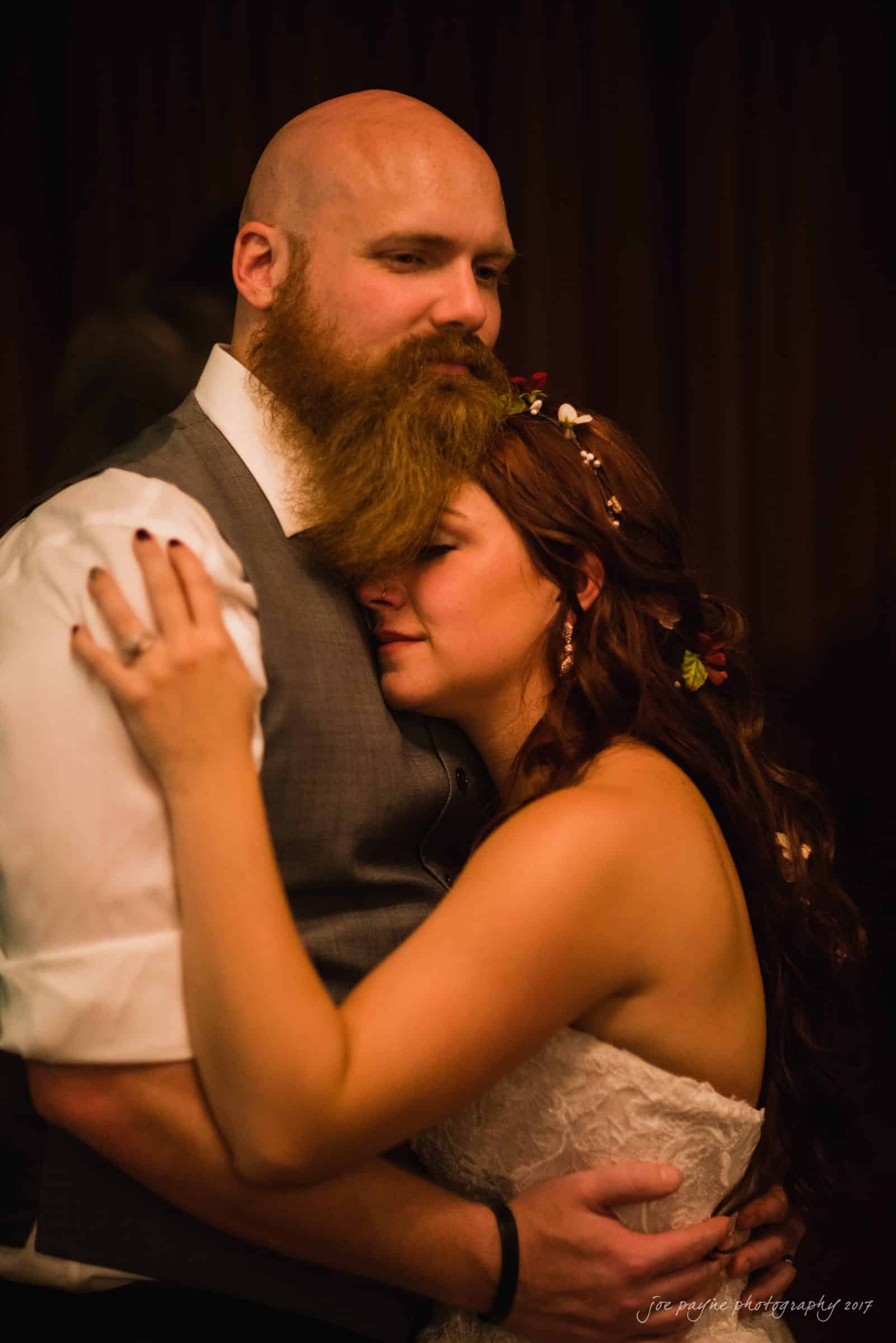 river landing country club wedding - megan & chad A couple embraces closely, sharing an intimate moment. The man has a beard and wears a vest, while the woman in a white dress rests her head on his chest. Both appear content and peaceful, enjoying the warmth and closeness of the moment.