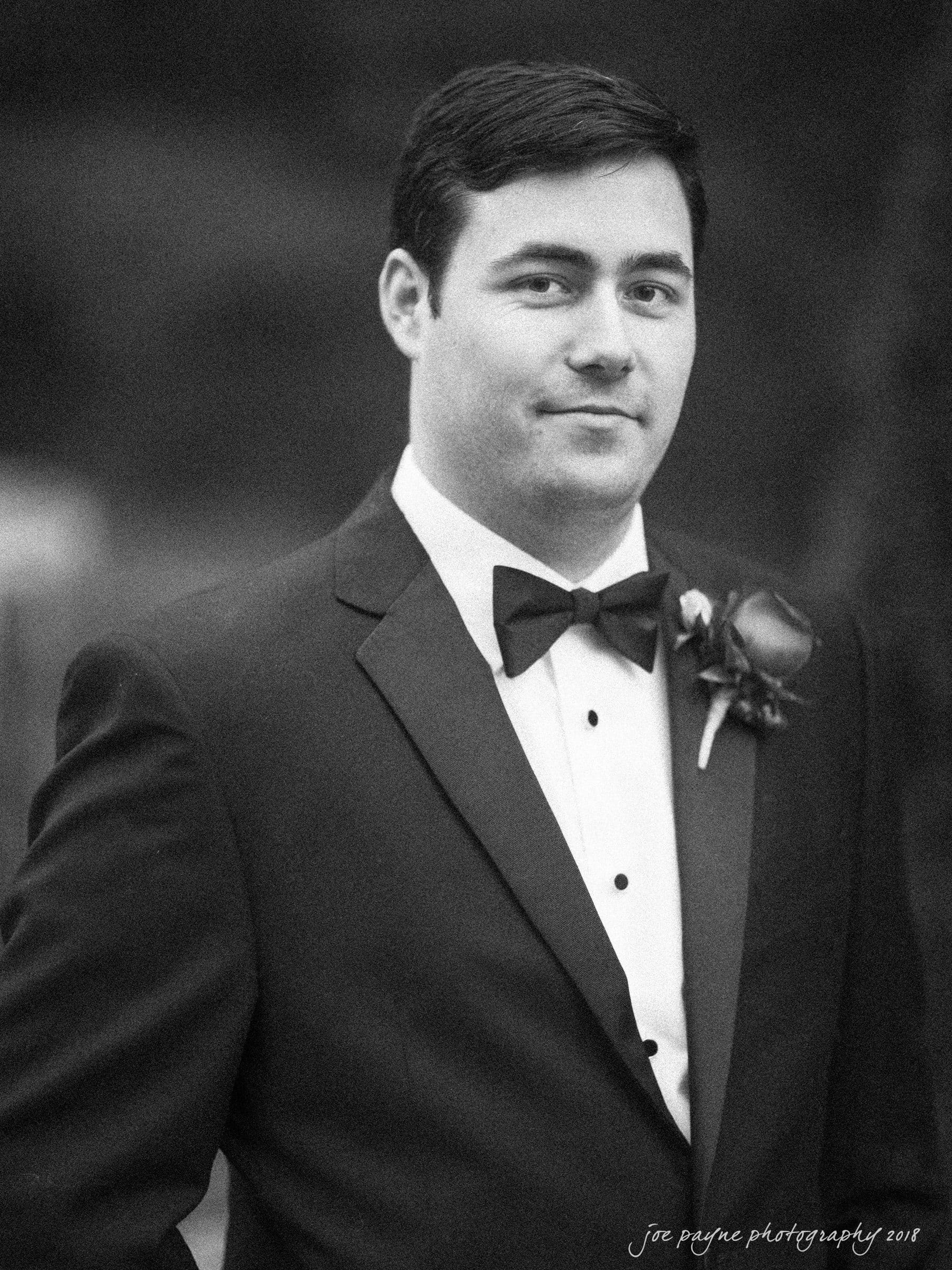 A man in a tuxedo with a bow tie and boutonniere poses outdoors. The photo has a soft, monochrome finish and is signed Joe Payne Photography 2011 in the corner.