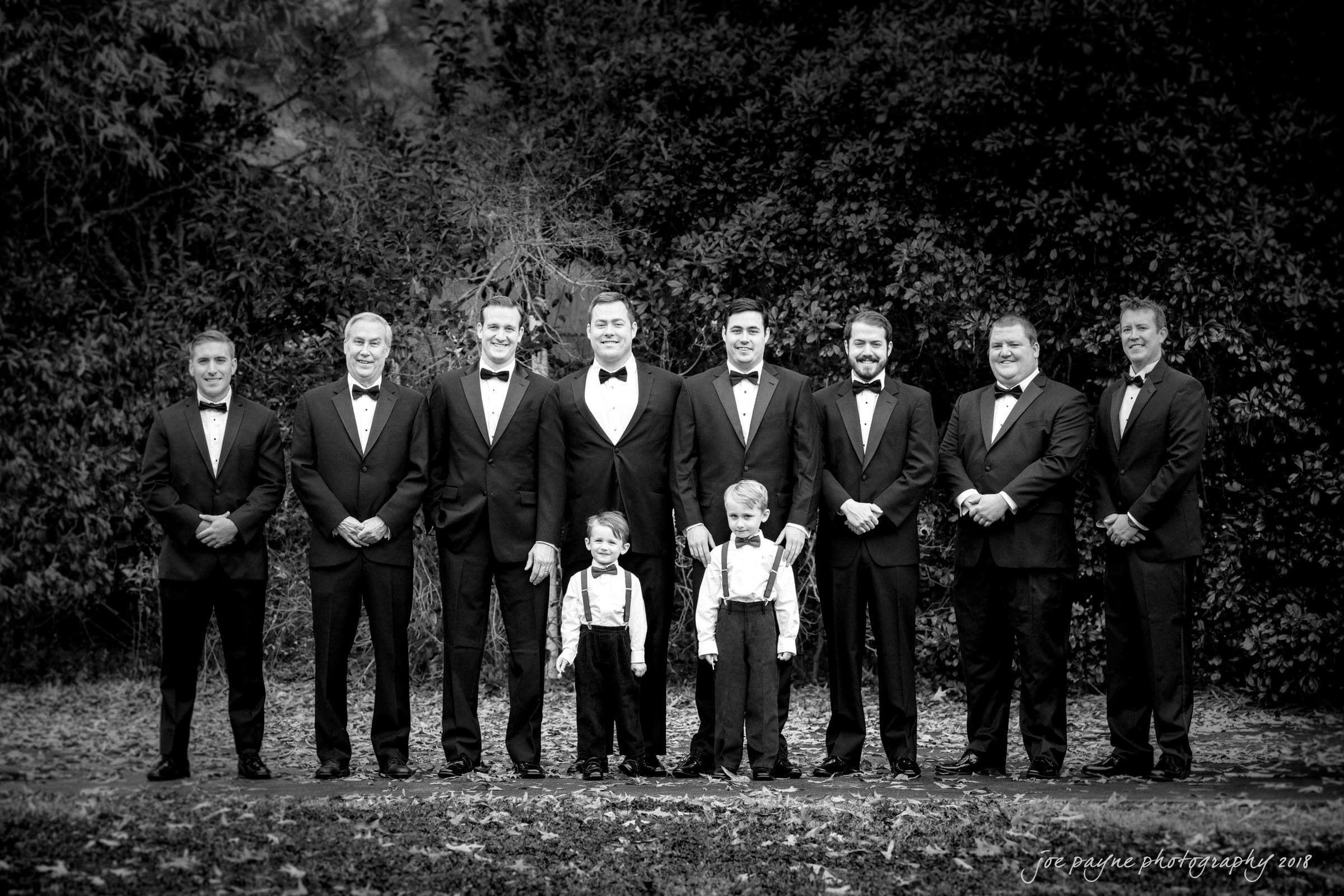 A group of nine men and two young boys in formal attire stand outdoors in a row. The men are wearing black suits and bow ties, while the boys are in suspenders and bow ties. The setting is a forested area with leaves on the ground.