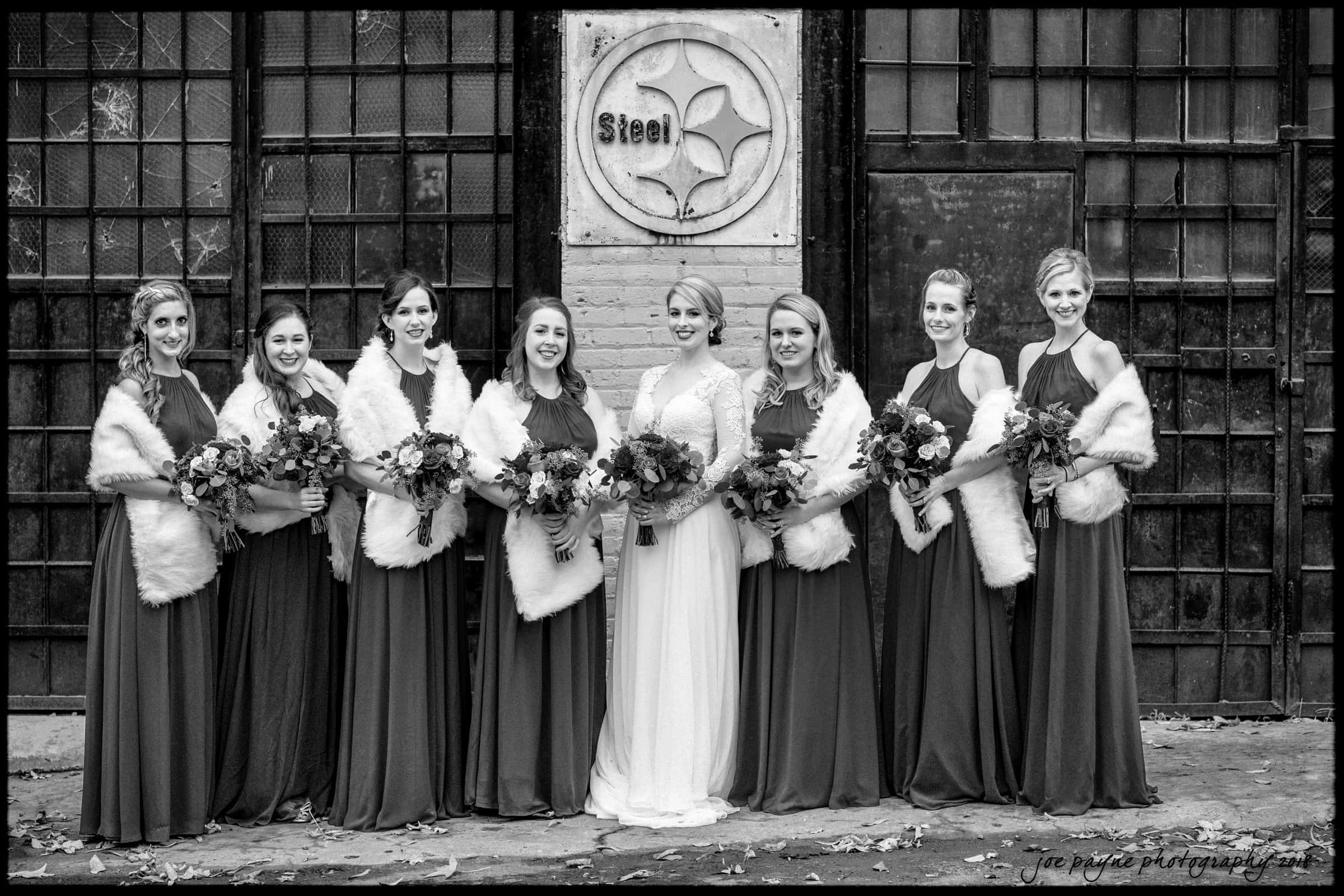 A black and white photo of a bride and seven bridesmaids standing in front of a rustic metal and brick wall. The bridesmaids wear dark gowns with white fur stoles and hold bouquets. The bride wears a white gown and also holds a bouquet.