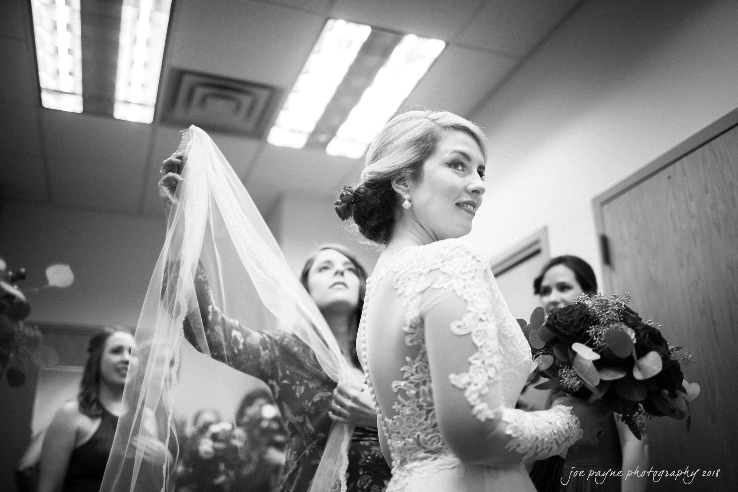 A bride in a lace gown holds a bouquet, looking to the side. Her veil is being adjusted by another woman. Two other bridesmaids are visible in the background, all inside a room with soft lighting.