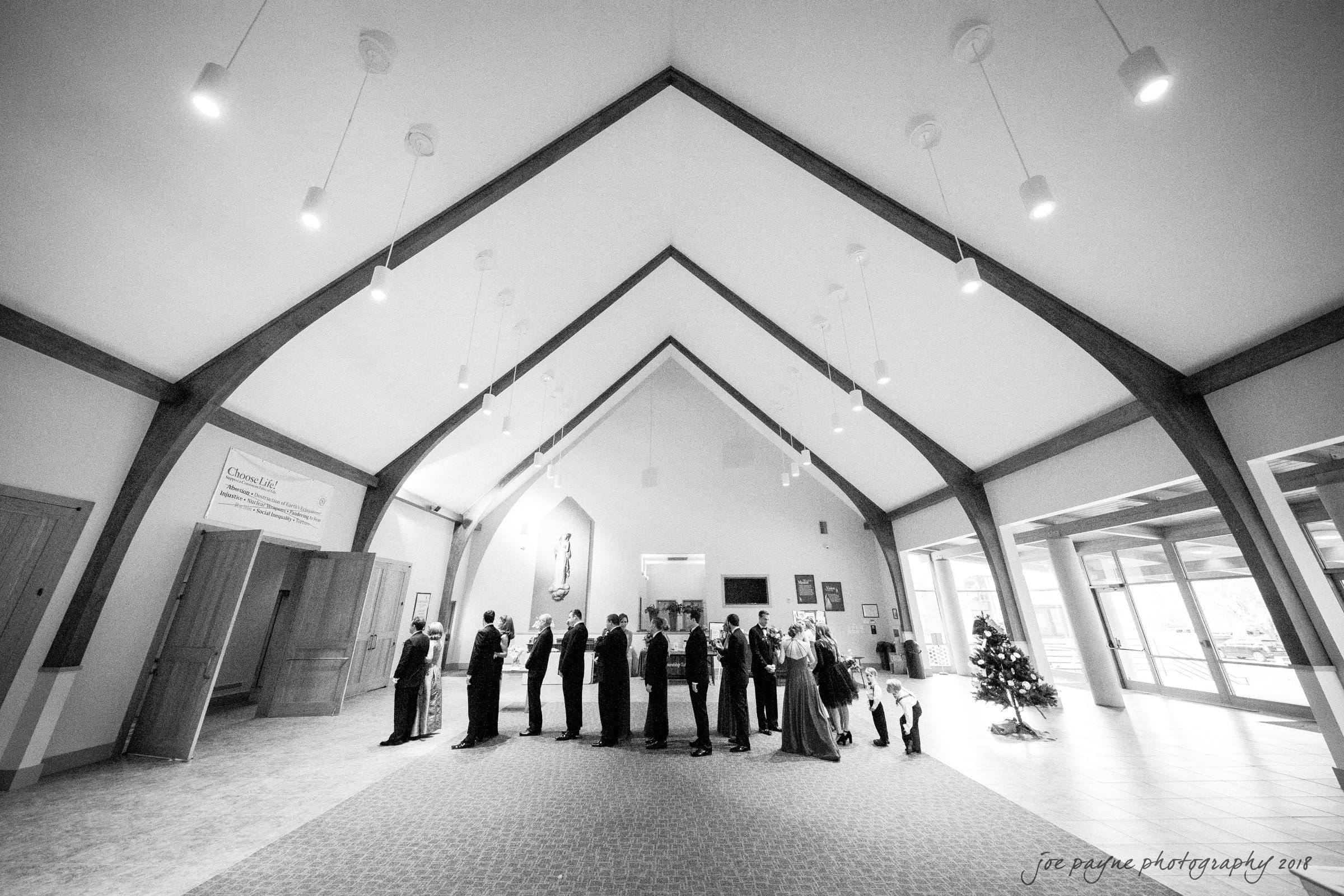 A black and white image of a group of people standing in a line inside a church with a high, triangular ceiling. They face the altar on the left. There are Christmas decorations, and light fixtures hang from the ceiling.