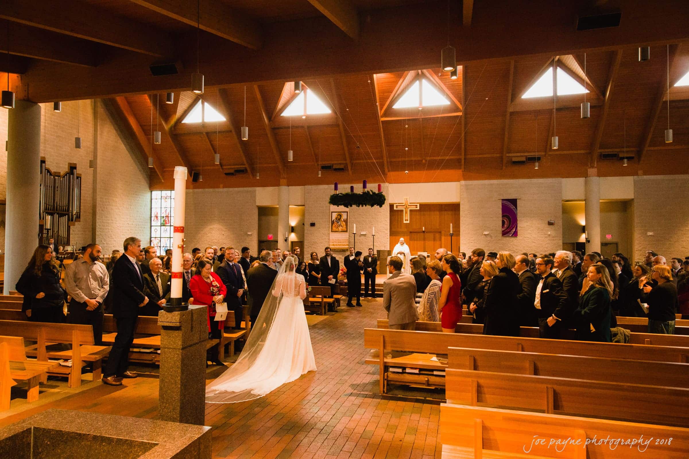 A bride with a long veil walks down the aisle of a church filled with seated guests. Sunlight streams through the windows, illuminating the wooden interior and high ceiling. An officiant stands at the altar adorned with greenery.