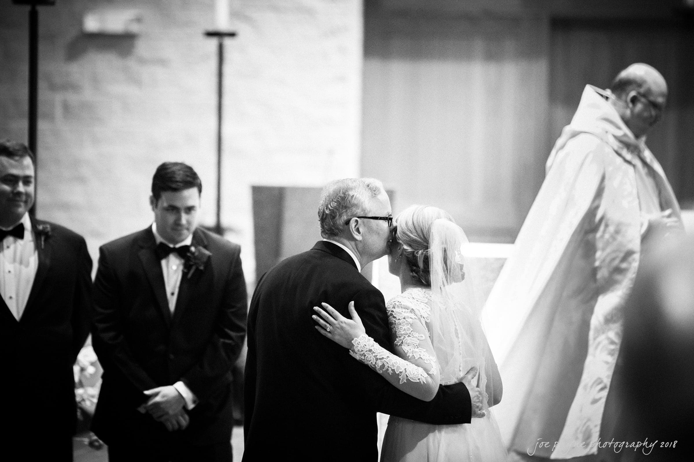 A black and white photo of a wedding ceremony where a man in a tuxedo kisses a bride wearing a lace gown on the cheek. They stand at the altar, with a priest in robes nearby and another man in a tuxedo observing.