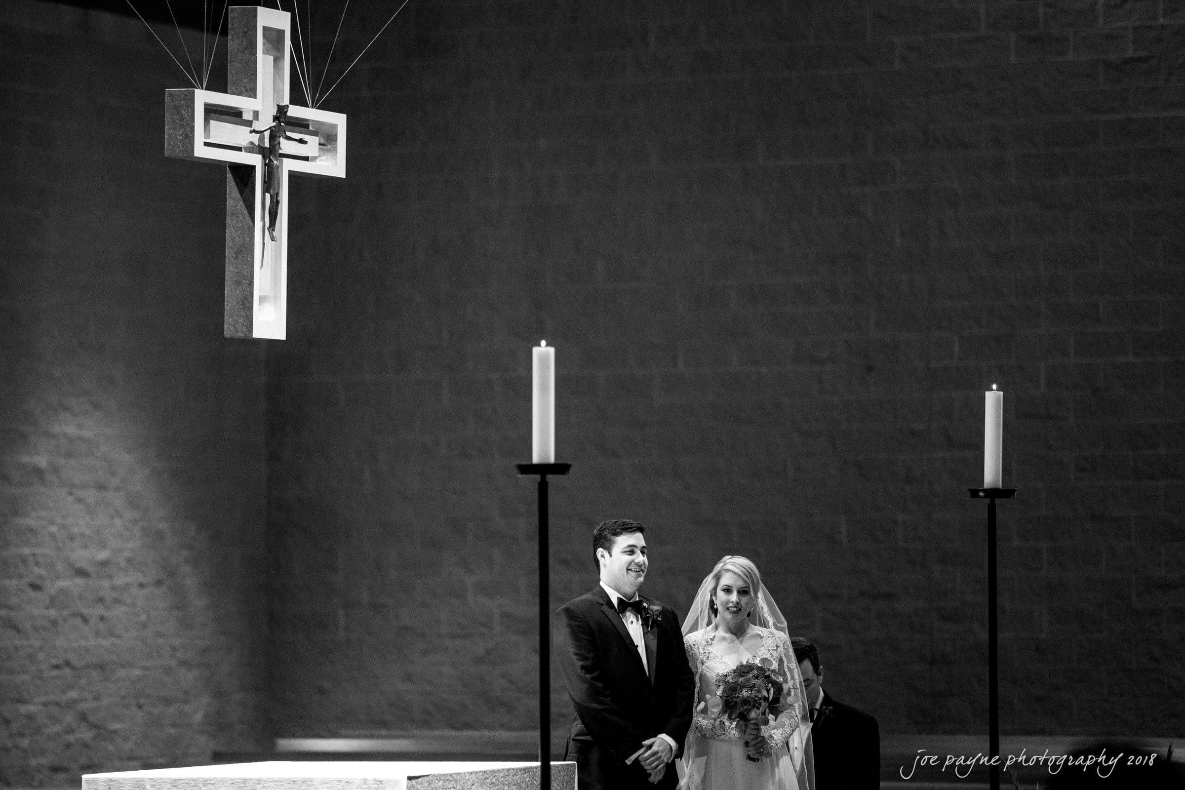 A bride and groom stand at an altar in a church. The couple is facing forward, with a large cross suspended above them. Two tall candles are visible on either side. The atmosphere is solemn and reverent.