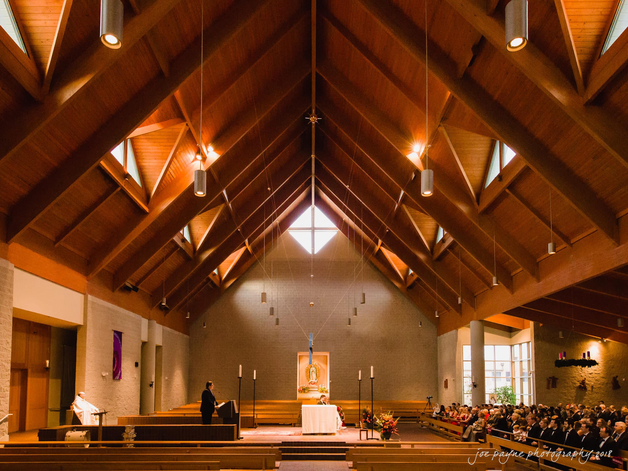 A spacious church interior with a wooden vaulted ceiling. The altar is centered with lit candles, and an image hangs above it. Many people are seated in pews on the right side, attending a ceremony.