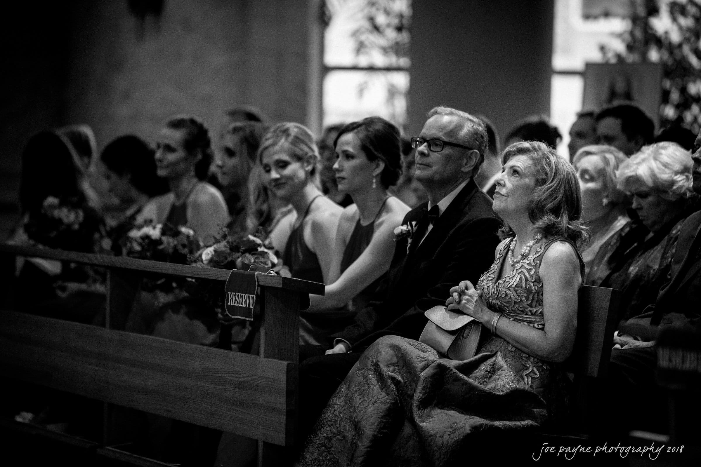 A black and white photo of an attentive audience seated on wooden benches in a dimly lit venue. The front row features elegantly dressed individuals, with a woman holding a program and others gazing ahead intently.