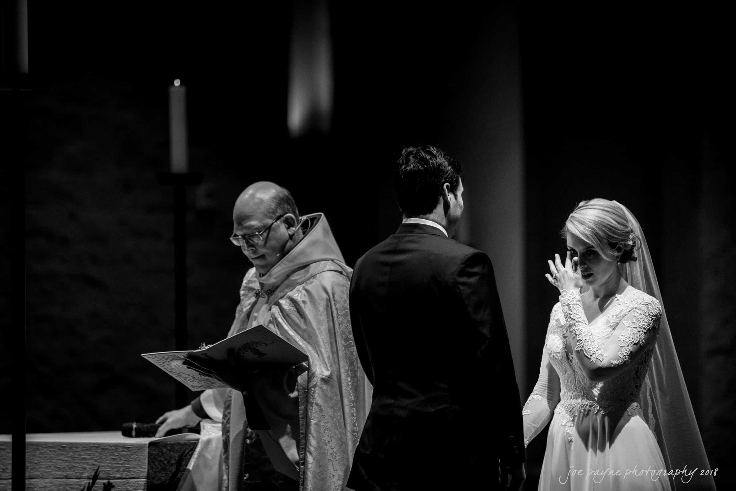 A black and white photo of a wedding ceremony. A bride in a lace gown wipes her eyes, while facing the groom in a suit. A priest in ceremonial attire stands in front of them, holding a book. Candles are visible in the background.
