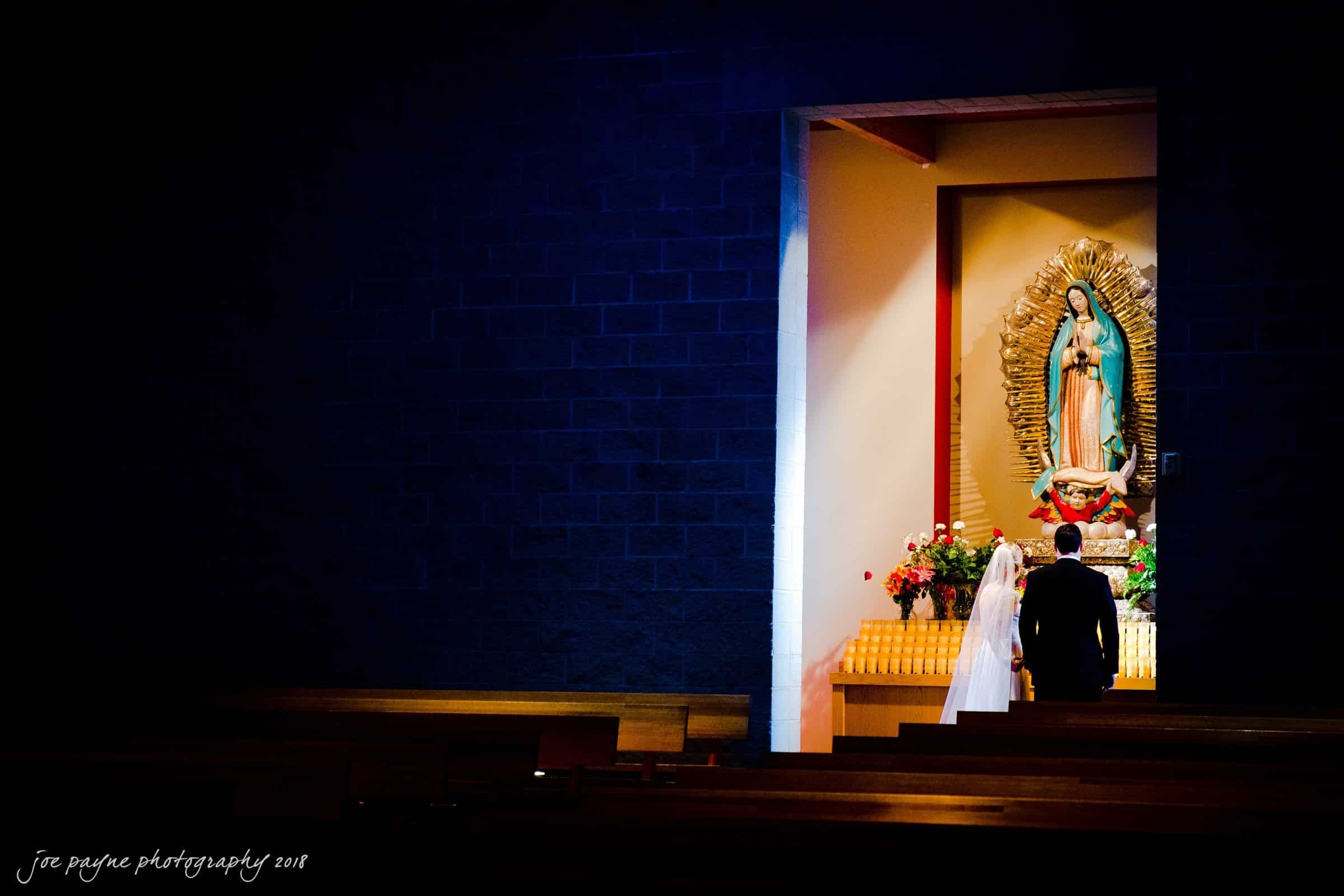 A couple stands at the altar in a dimly lit church, facing a statue of the Virgin Mary adorned with flowers. The bride is in a white dress and veil, and the groom is in a dark suit. The scene is tranquil and reverent.