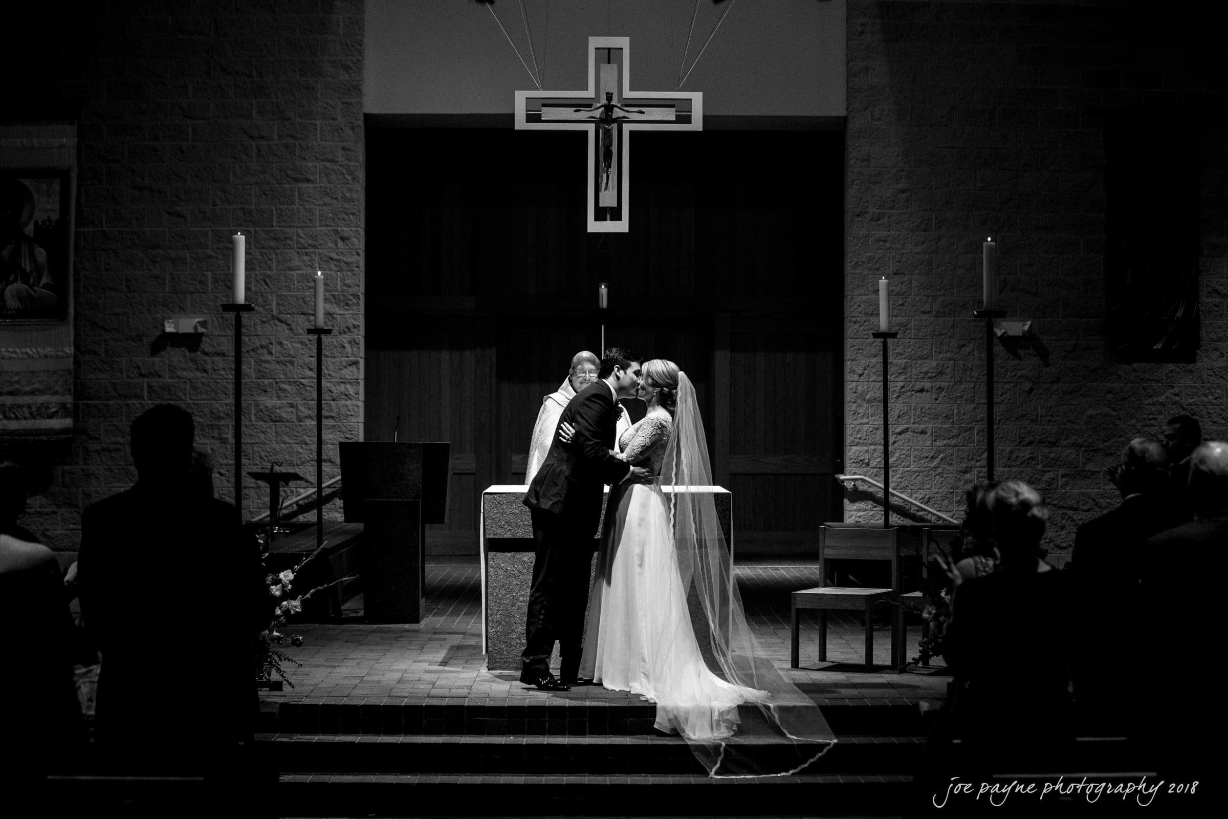 A couple kisses at the altar during their wedding ceremony in a church. The bride is in a white gown and veil, and the groom is in a suit. A large cross hangs above them, and guests are seated in the pews. The atmosphere is solemn and intimate.