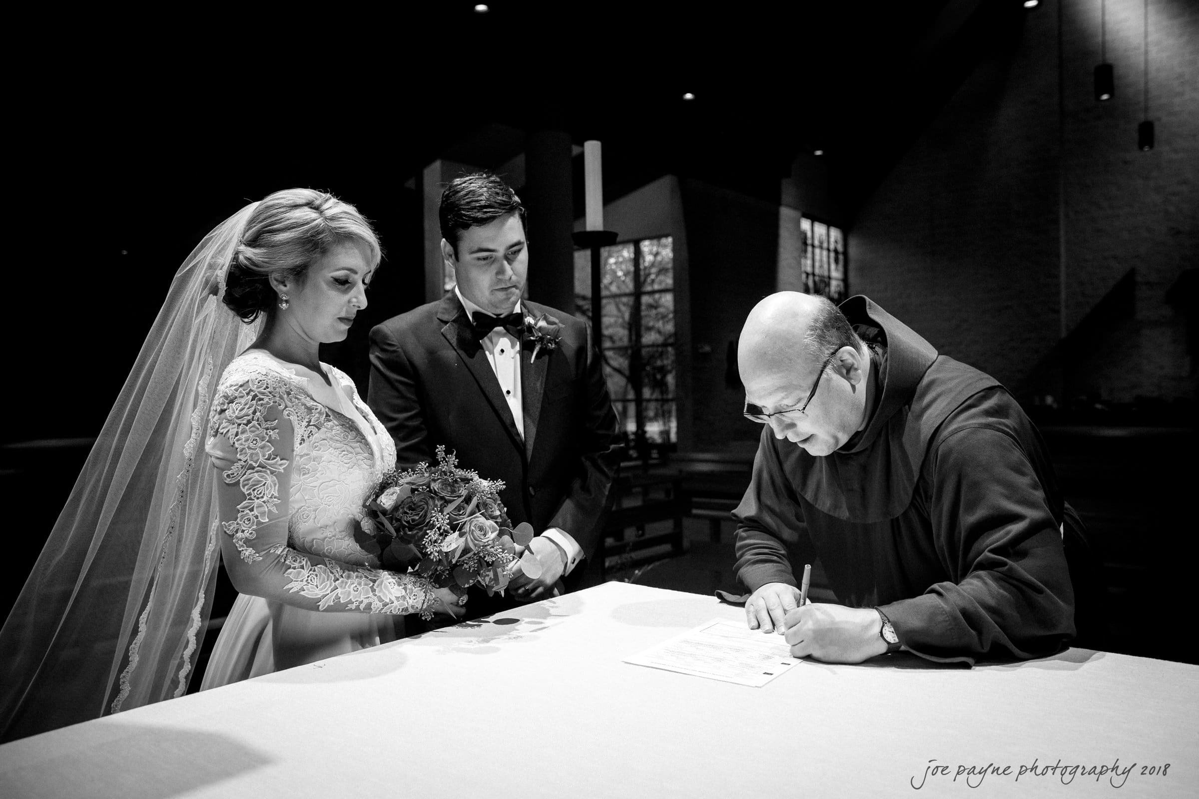 A bride and groom stand before a clergyman who is signing a document on an altar in a dimly lit church. The bride holds a bouquet and wears a lace wedding dress, while the groom is in a tuxedo. The scene is solemn and intimate.