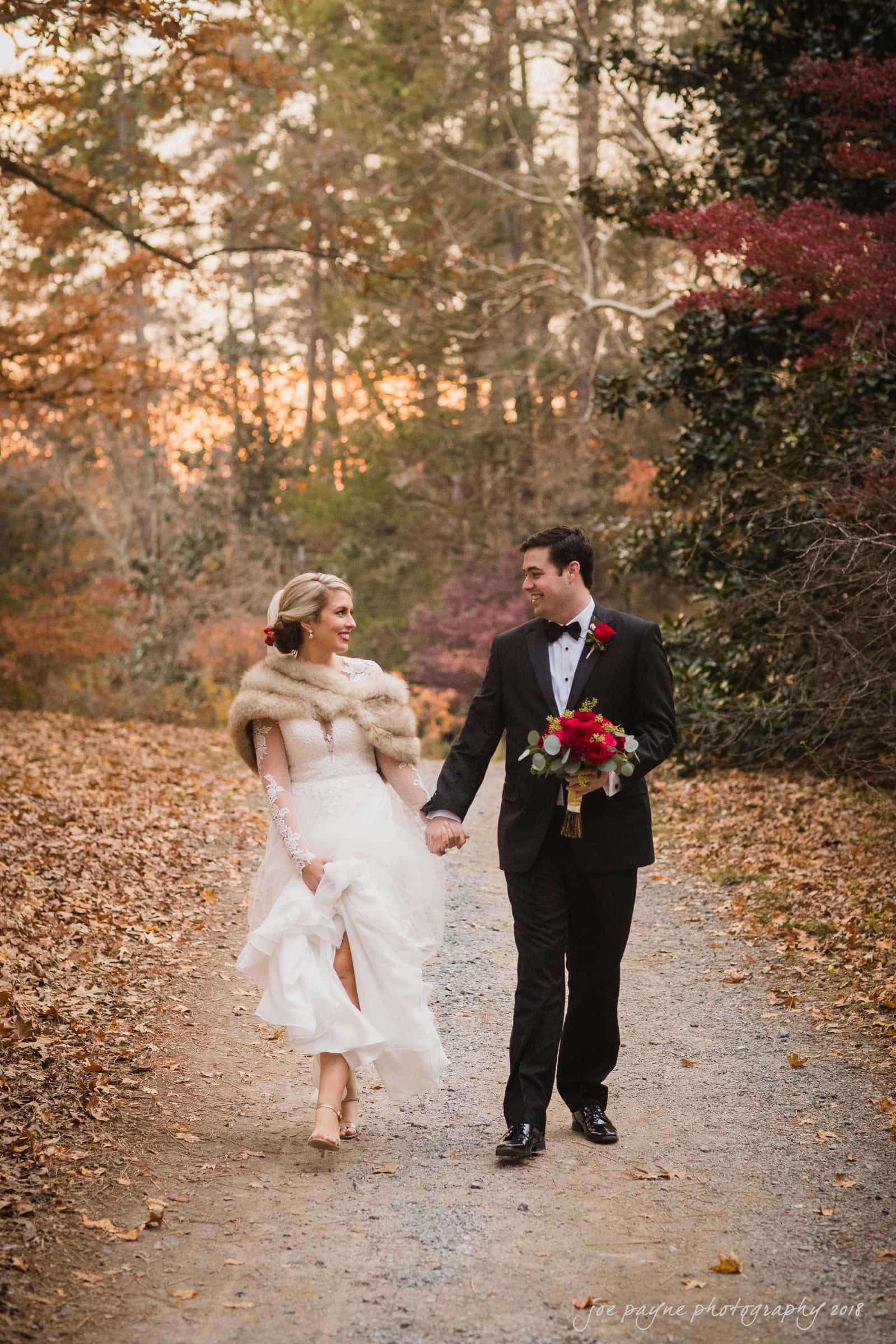 A bride and groom walk hand-in-hand down a leaf-covered path in autumn. The bride wears a white gown with a fur shawl, and the groom is in a black suit holding a bouquet. Trees with fall foliage frame the background.