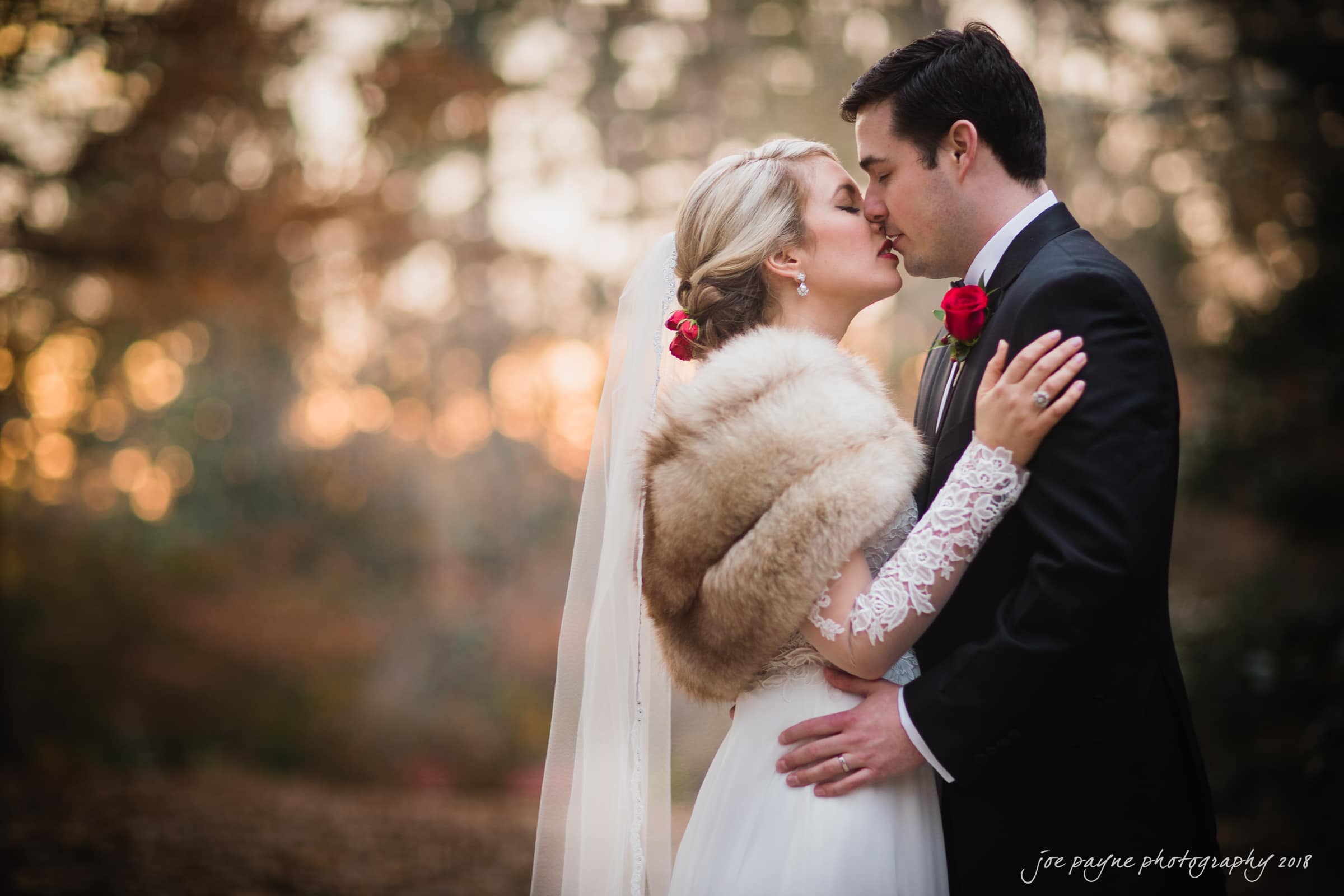 A bride and groom share a tender moment outdoors. The bride, wearing a lace gown and fur stole, embraces the groom, who is in a dark suit with a red rose boutonniere. They are surrounded by softly blurred trees in autumn hues.