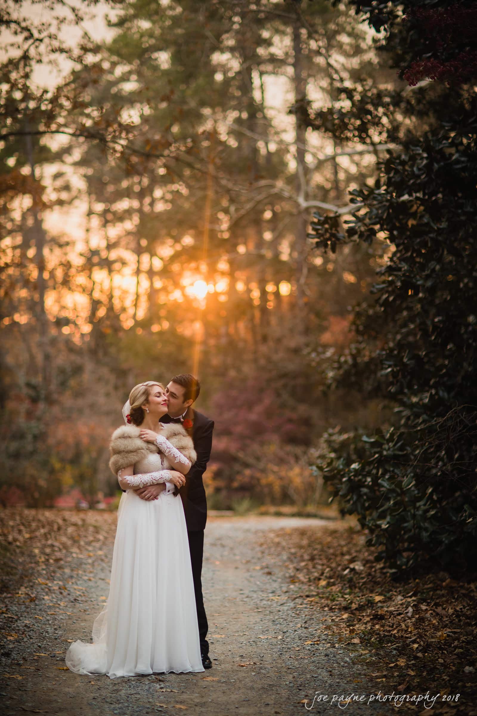 A couple in wedding attire stands on a forest path at sunset. The bride, wearing a white gown and fur shawl, is embraced by the groom in a suit. Warm sunlight filters through the trees, creating a romantic atmosphere.